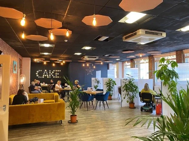 People sitting at tables in a cozy cafe with a dark ceiling, decorative lighting, and green potted plants near large windows.