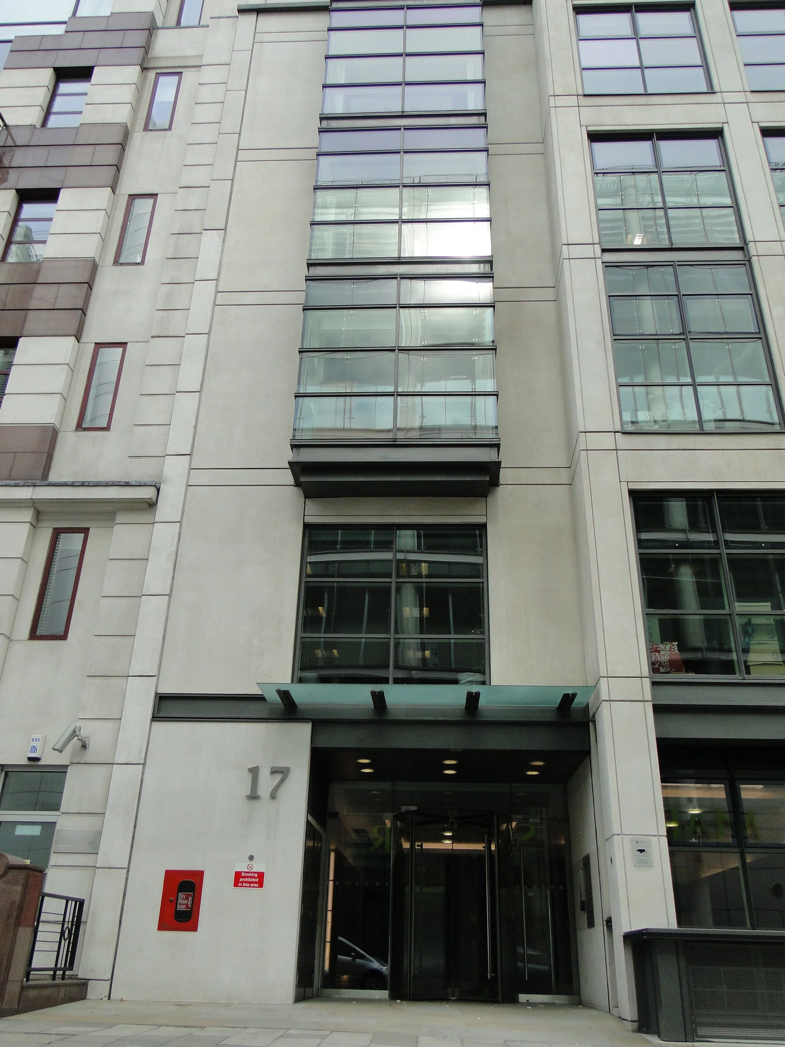 The entrance to a modern multi-story building with the number 17 displayed on the wall, glass doors, and large windows reflecting the city surroundings.