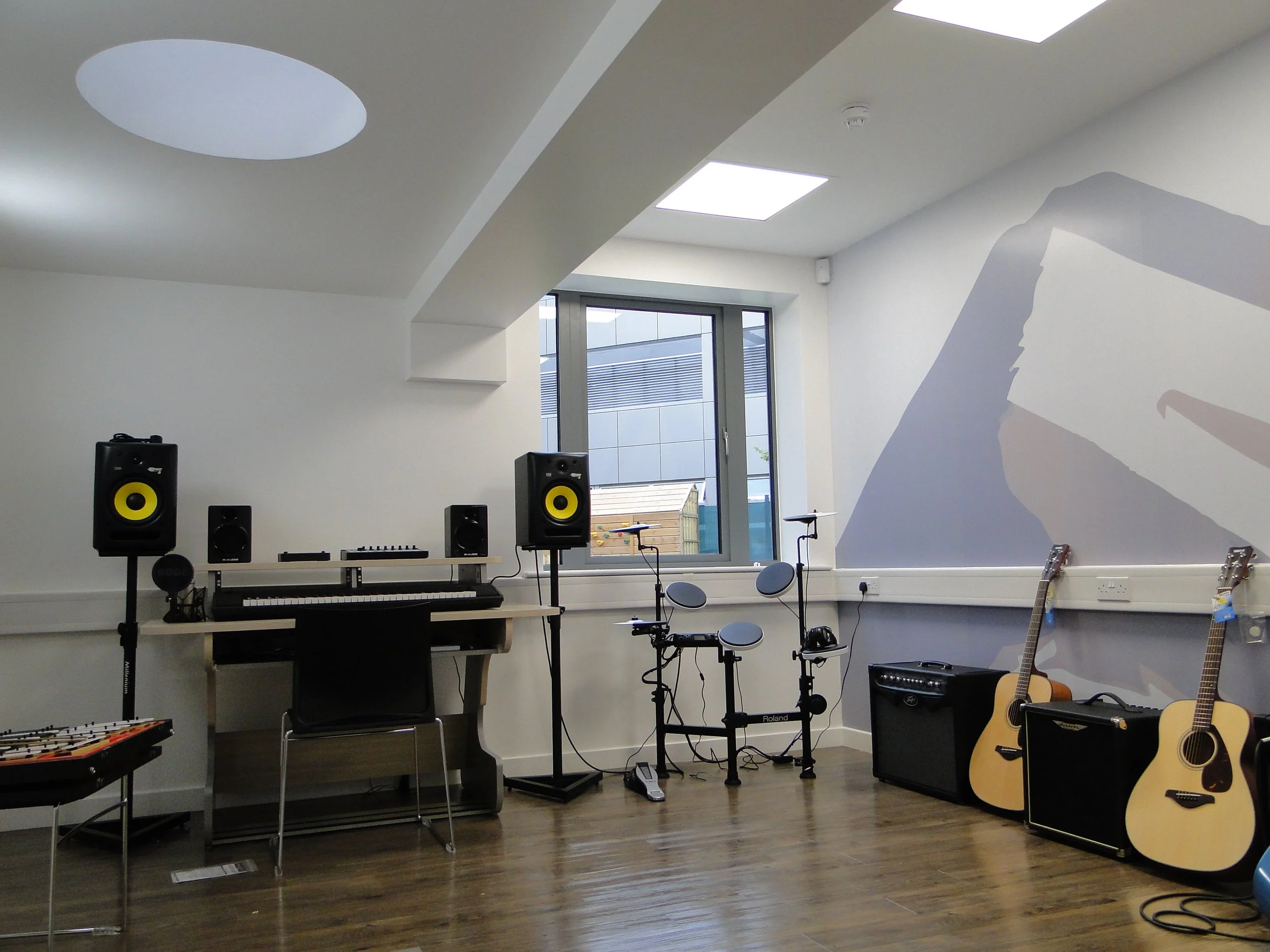 Music practice room with guitars, microphones, speakers, digital drum set, and a keyboard, with wooden flooring and white walls featuring a large abstract mural.