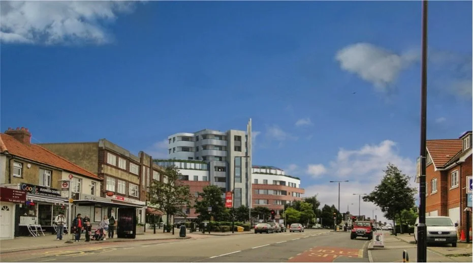 A city street with shops on the left, cars on the road, and a modern multi-story building in the background under a blue sky with a few clouds.