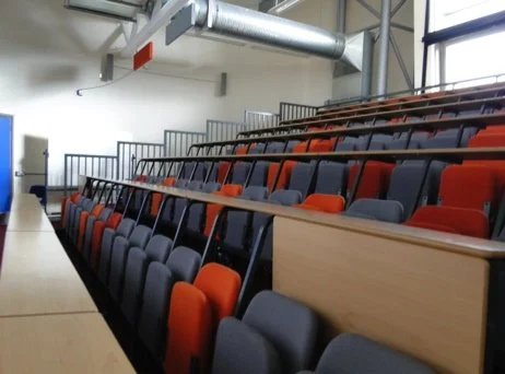 Empty lecture hall with tiered seating, gray and orange chairs, and a wooden desk at the front.