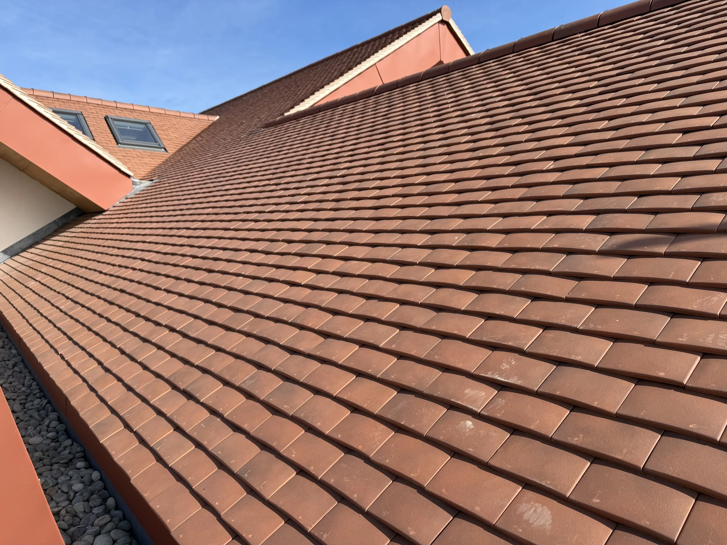 Close-up view of a roof with reddish-brown tiles, showing a section of the roof and two skylights, under a clear blue sky.