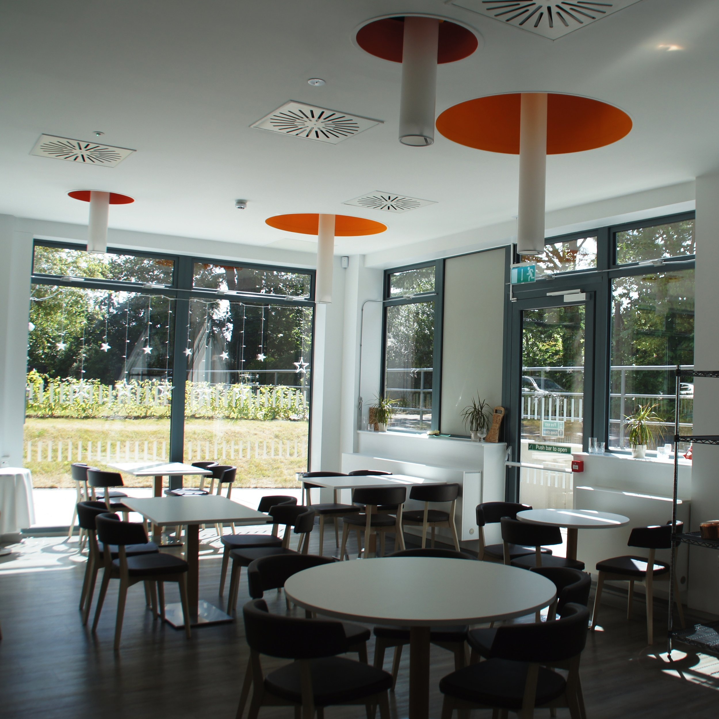 Interior of a modern cafe or restaurant with round and square tables, black chairs, large windows, and orange and white ceiling decorations.
