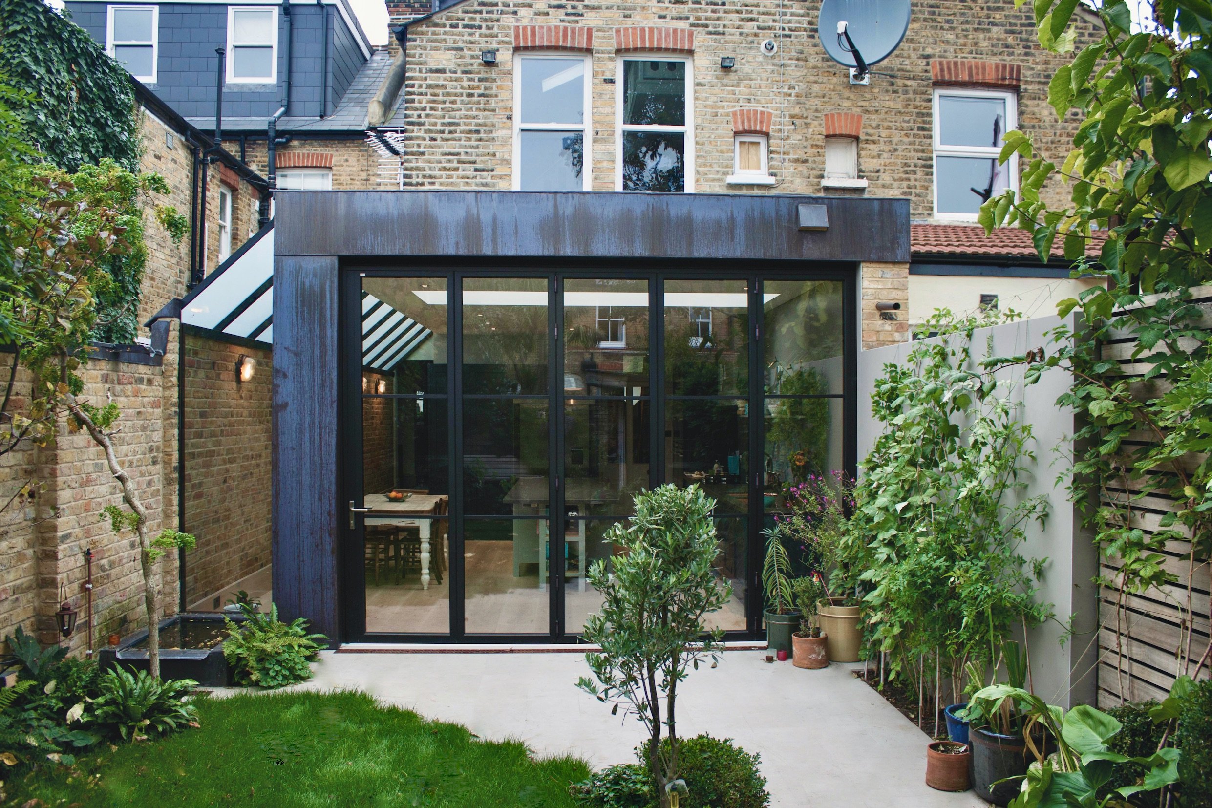 Backyard garden featuring a modern glass-walled extension with potted plants, surrounding brick and wooden fences, small trees, and a lush green lawn.