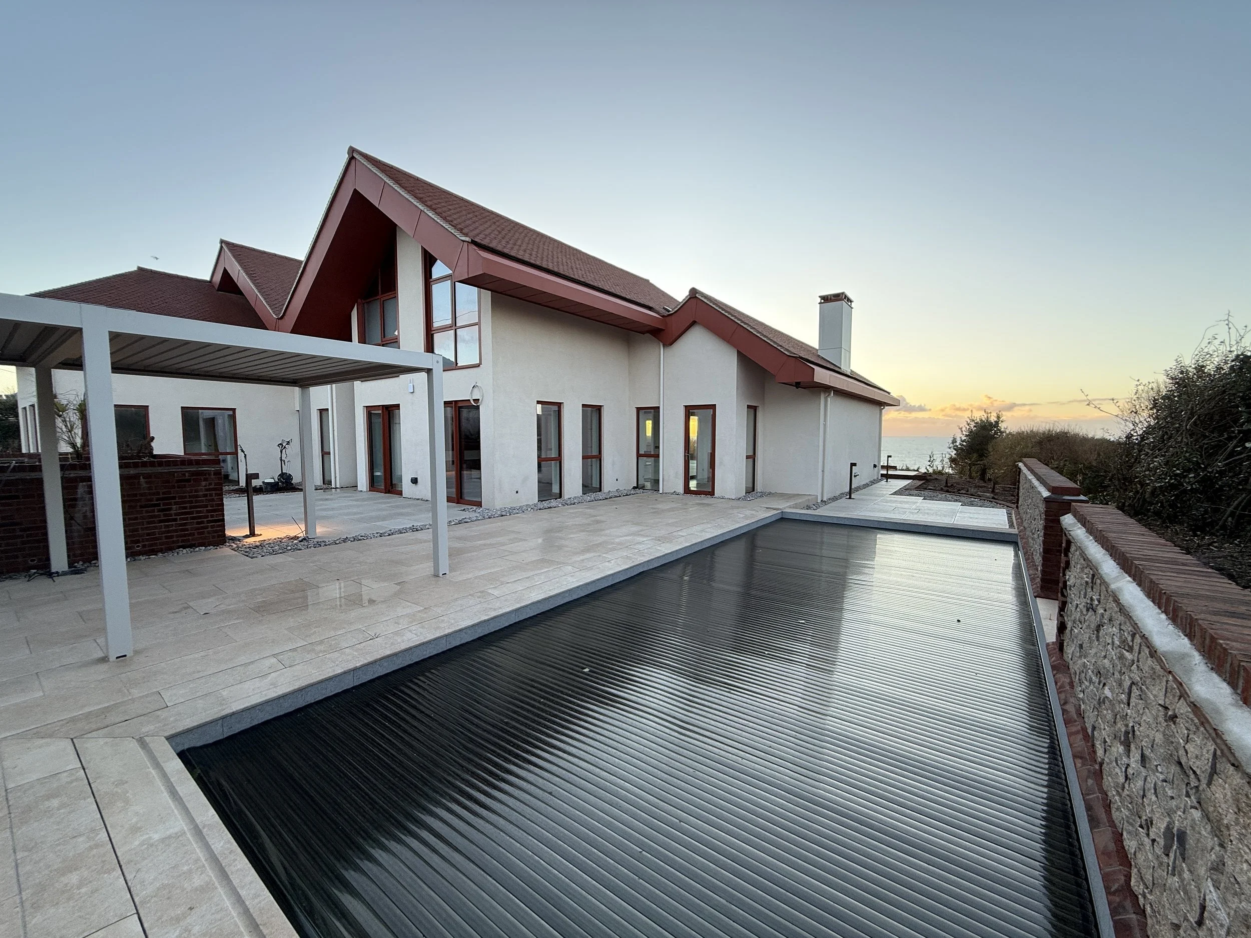 Modern house with red roofs, large glass doors, a patio area, and a black roll-up swimming pool cover at sunset, overlooking the ocean.