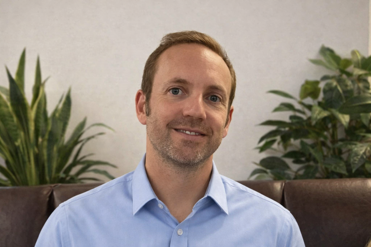 A man with light brown hair and a short beard, wearing a blue shirt, sitting in front of a light-colored wall.