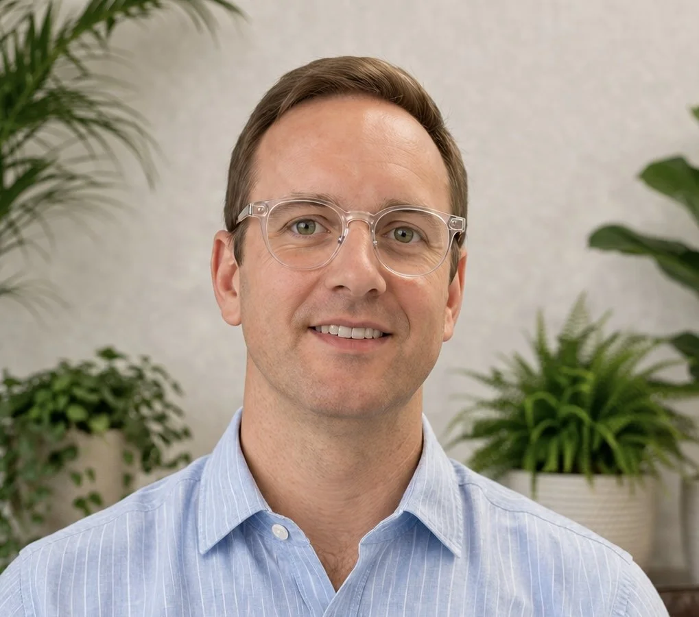 A man with light brown hair and a short beard, wearing a blue shirt, sitting in front of a light-colored wall.