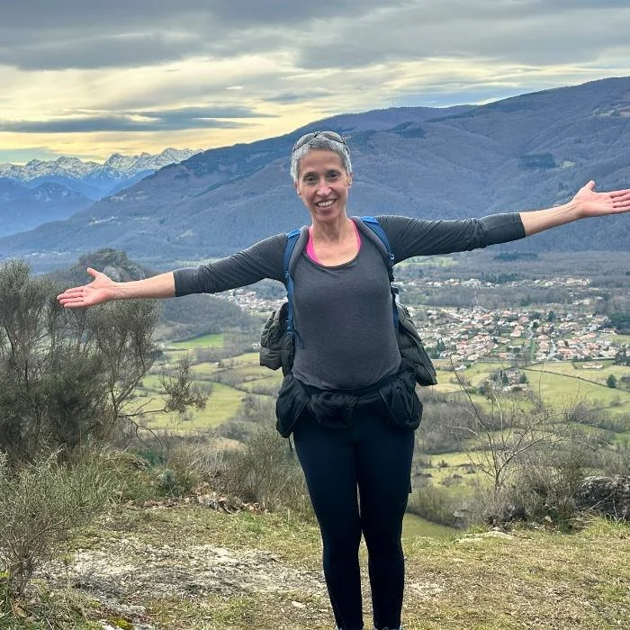 Middle aged woman with short grey hair staining with her arms outstreatched with mountains in the background.