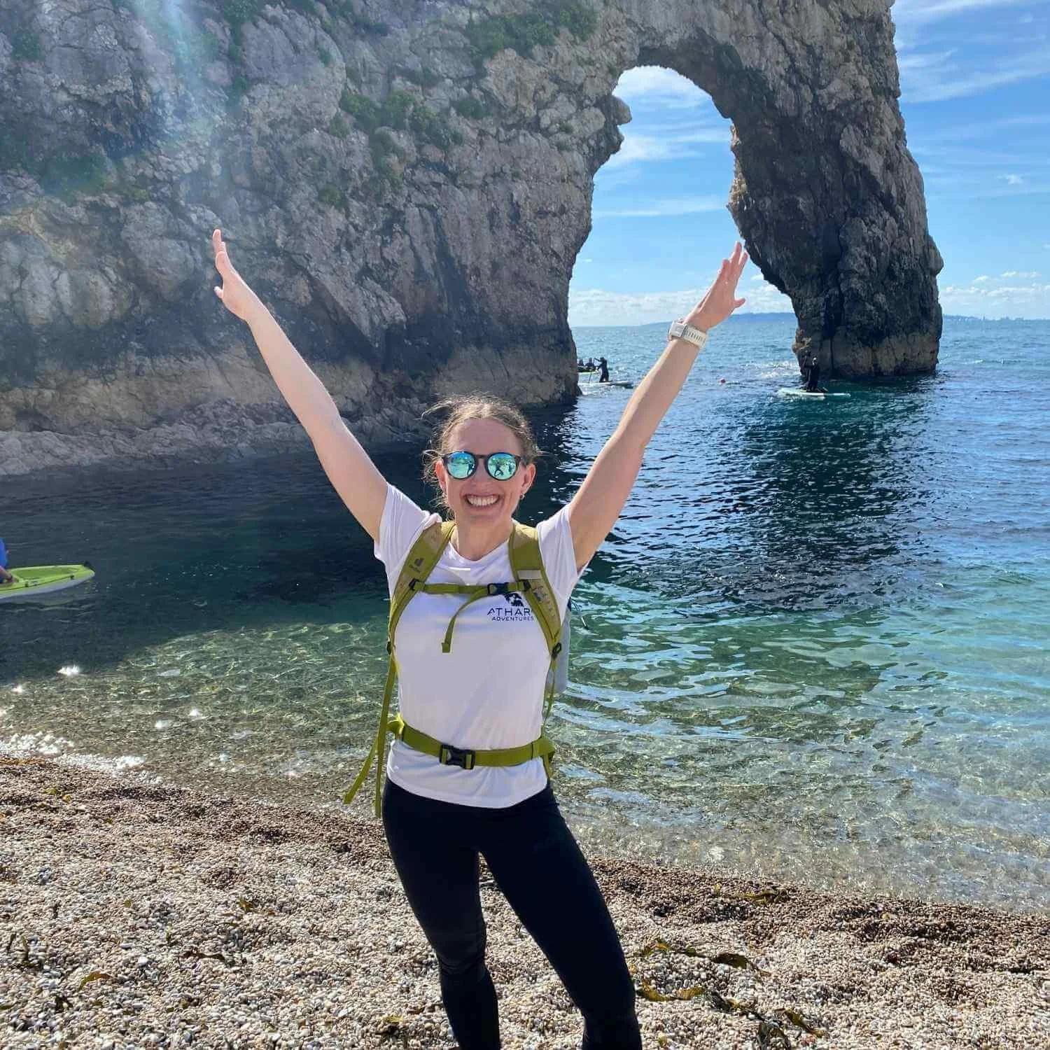 Sarah standing on a rocky shoreline with arms raised, wearing an Athara Adventures shirt and backpack, with clear blue water and cliffs behind her.