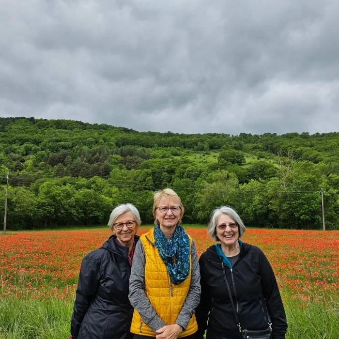 Three smiling women in their 70s standing in a field of red poppies.
