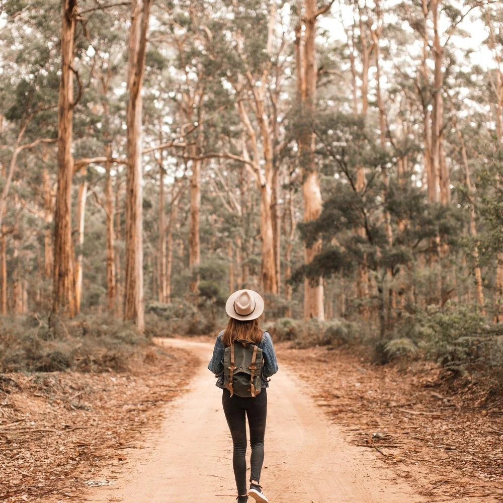 Woman walking down a tree-lined pathway wearing a hat and backpack.