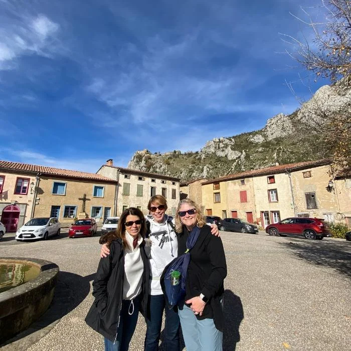 Three middle aged women smiling and embracing with a french village in the background.