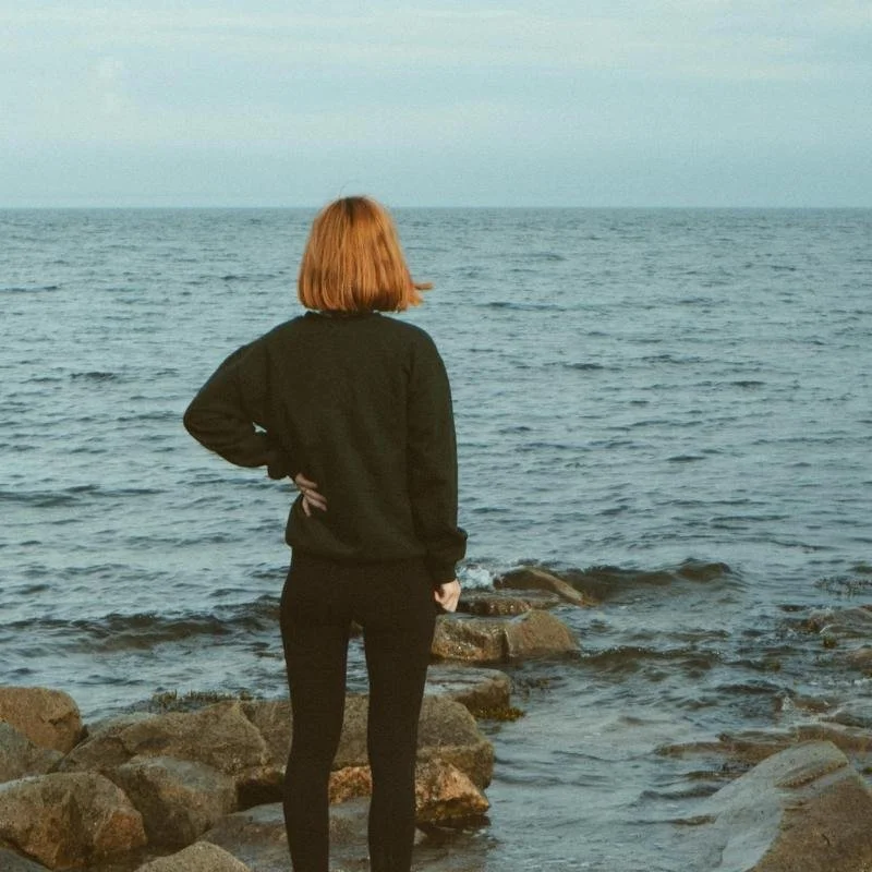 Red haired woman standing with hand on hip at the edge of the ocean.