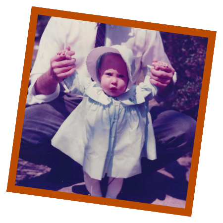 Baby Sarah standing outdoors with support, wearing a light dress and bonnet in a childhood photograph.