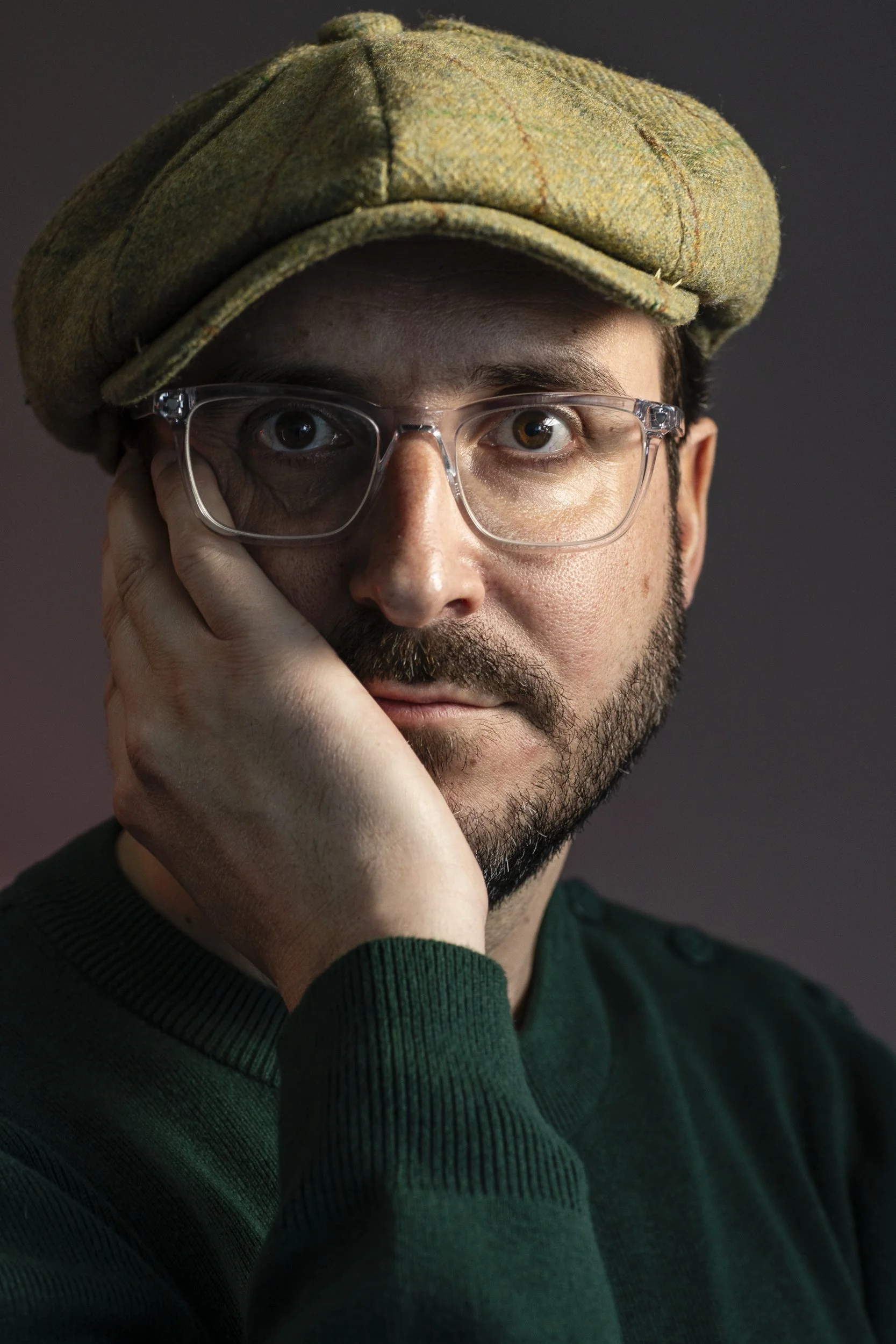 A close-up portrait of a man with brown eyes, wearing clear glasses and a green sweater, with a brown checkered cap on his head. He rests his chin on his hand, looking directly at the camera.