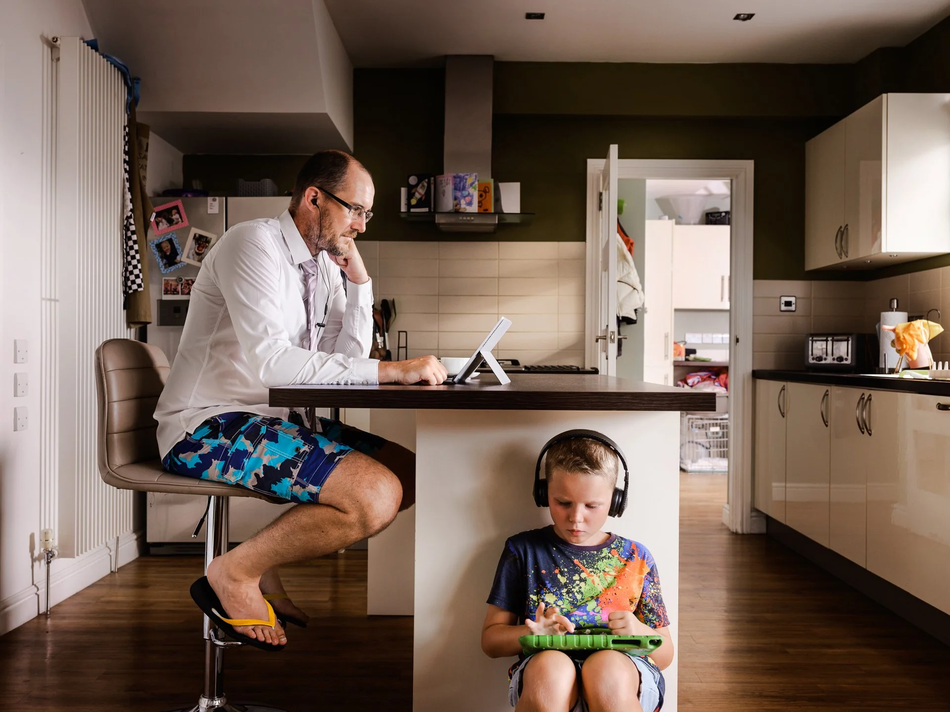 A man in a white shirt and blue camouflage shorts sits on a tall chair in the kitchen, using a tablet with earphones. A young boy in a colorful shirt and shorts, wearing headphones, sits under the kitchen counter, focused on a green handheld gaming device.