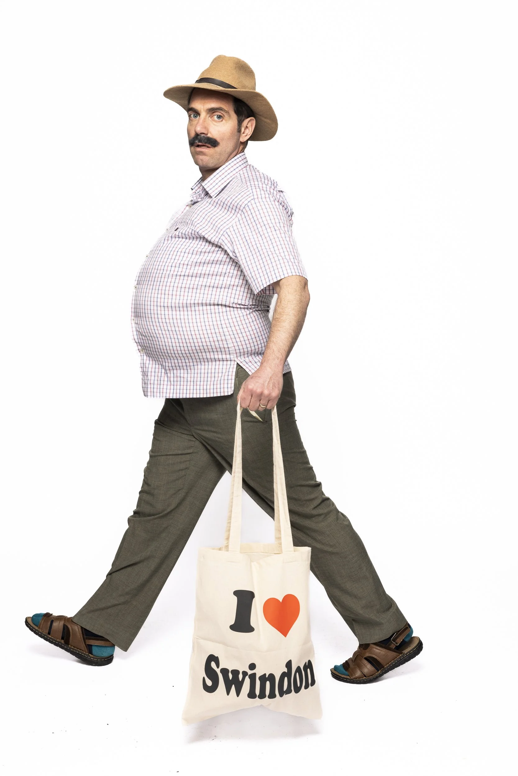 A man with a mustache wearing a hat, plaid shirt, and brown pants, standing with legs apart and holding a tote bag that says 'I ❤️ Swindon'.