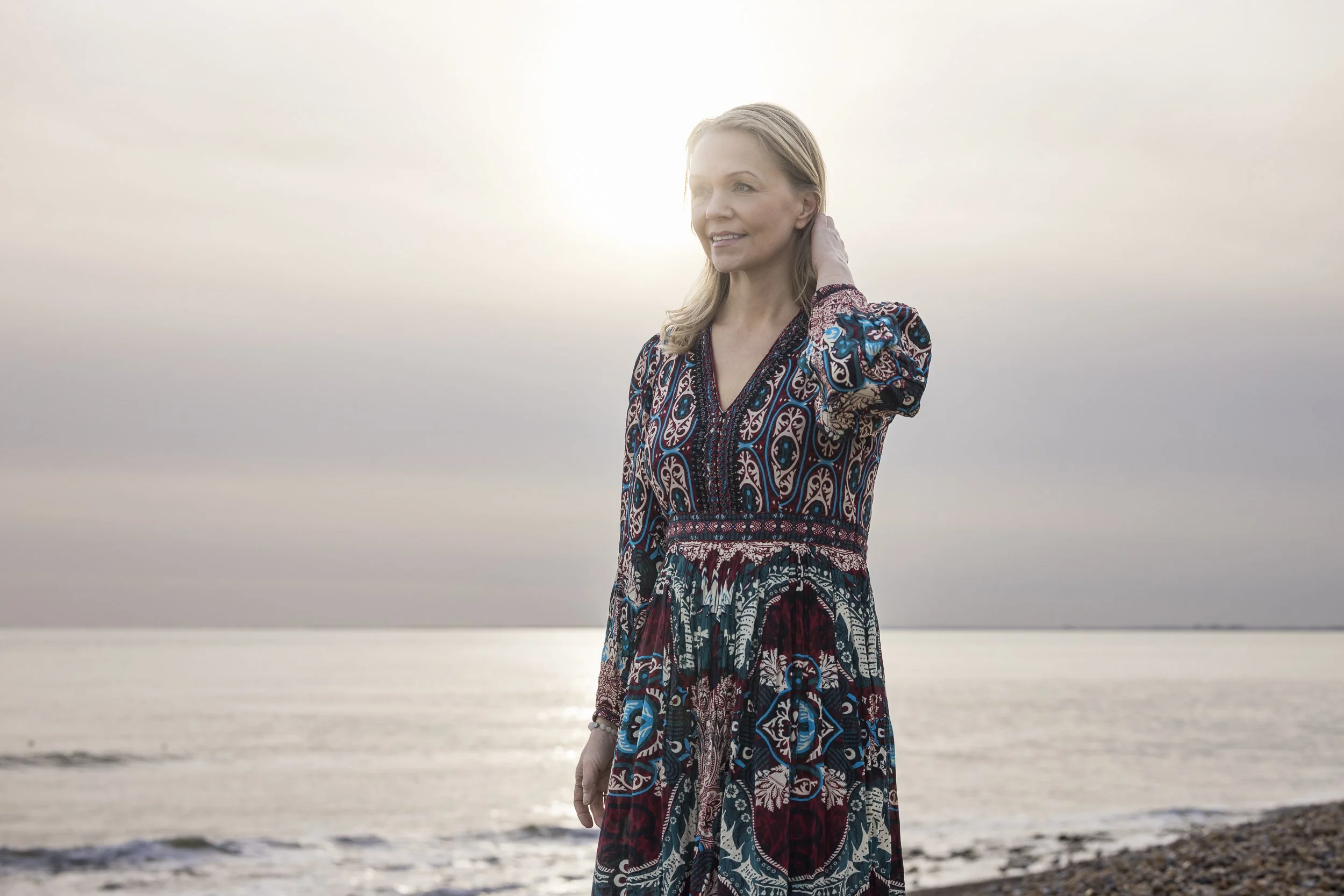 A woman with blonde hair wearing a colorful patterned dress standing on a beach in front of the ocean during sunset.