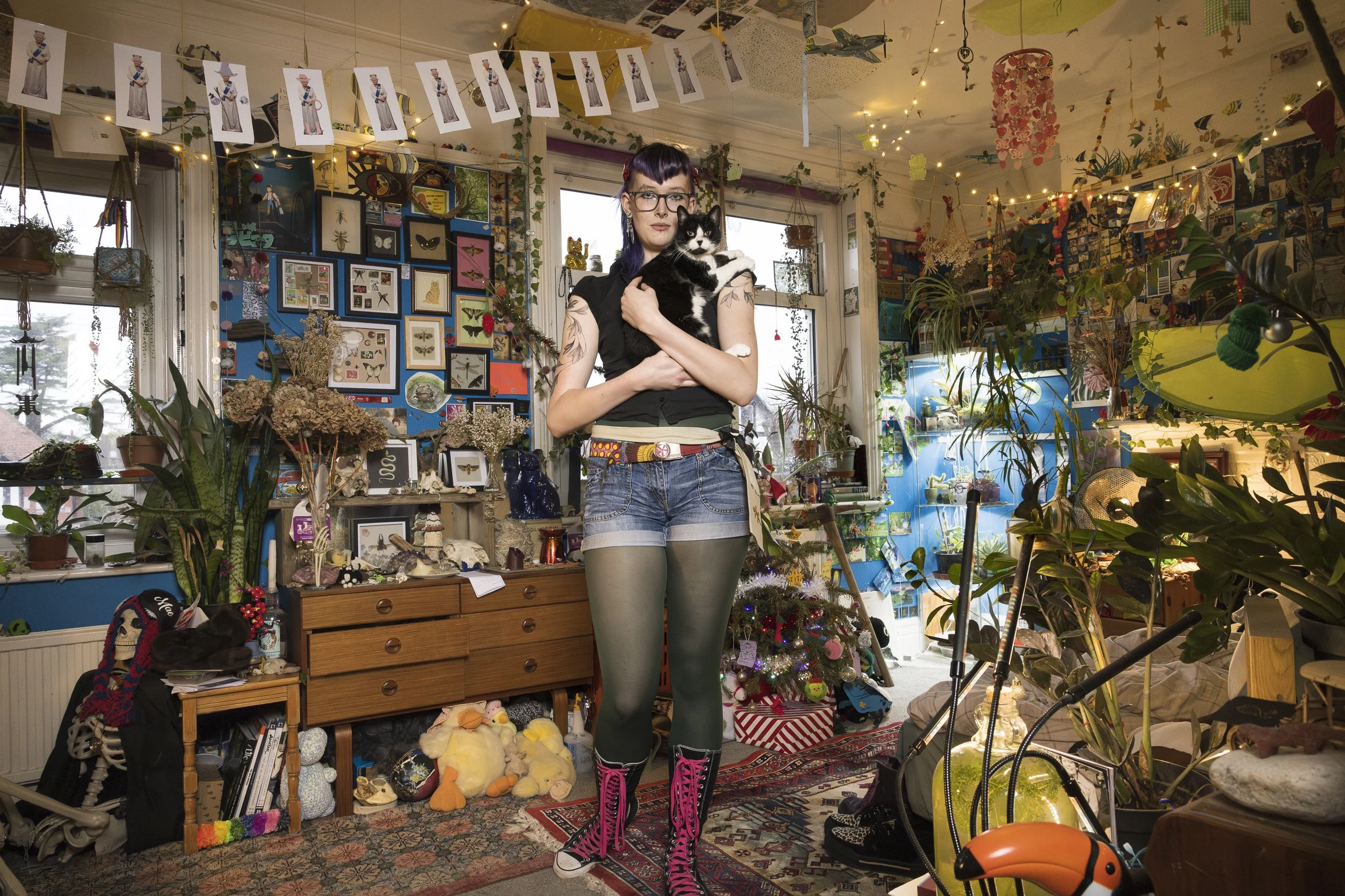 A woman with purple hair and tattoos on her arms is holding a black and white cat. They are in a cluttered room decorated with string lights, framed pictures of insects, and various plants. The room has a cozy, artistic vibe with a Christmas tree and assorted toys and objects around.