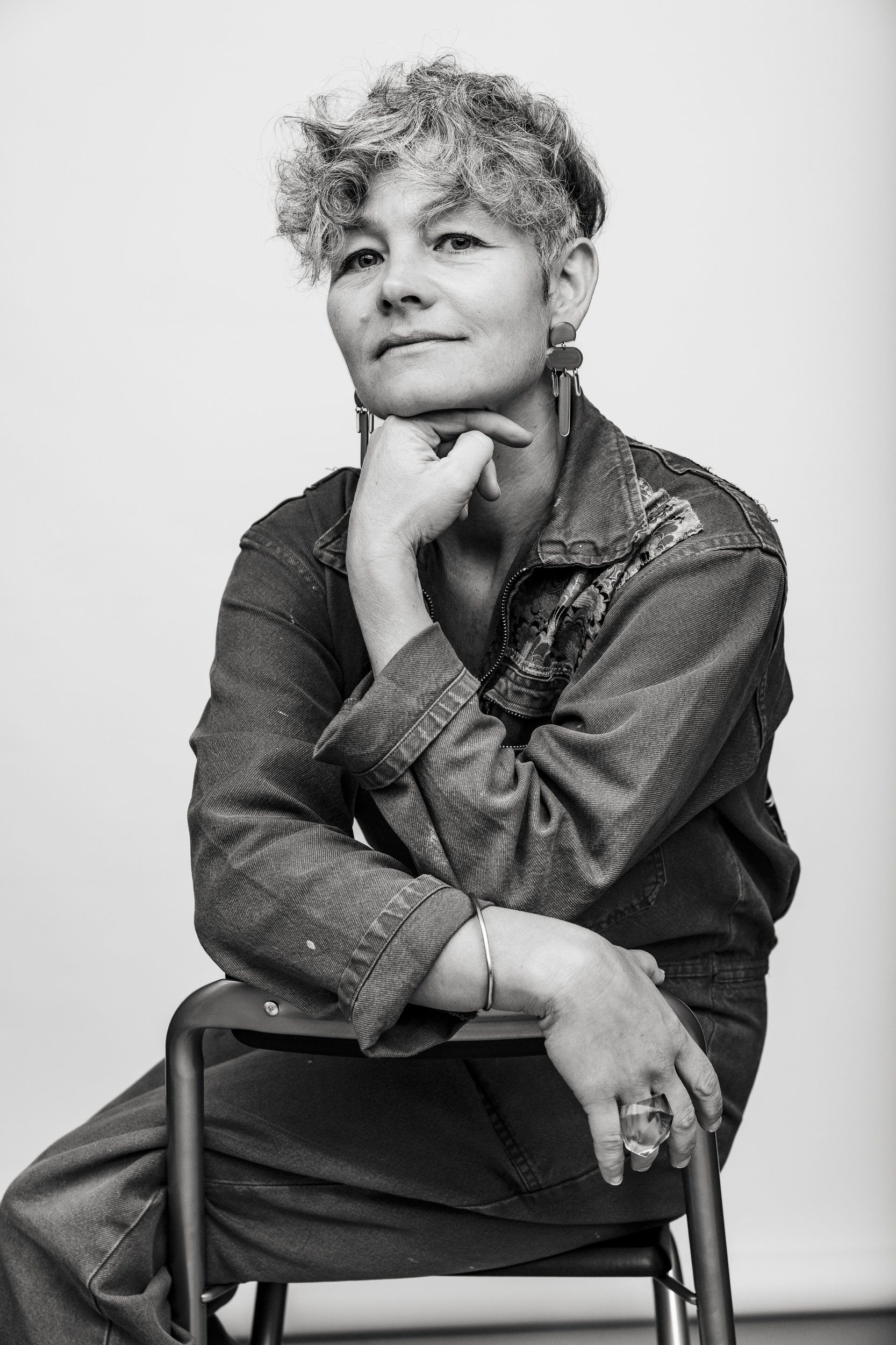 Black and white portrait of a person sitting on a chair, resting their chin on their hand, wearing earrings and casual clothing with a textured jacket.