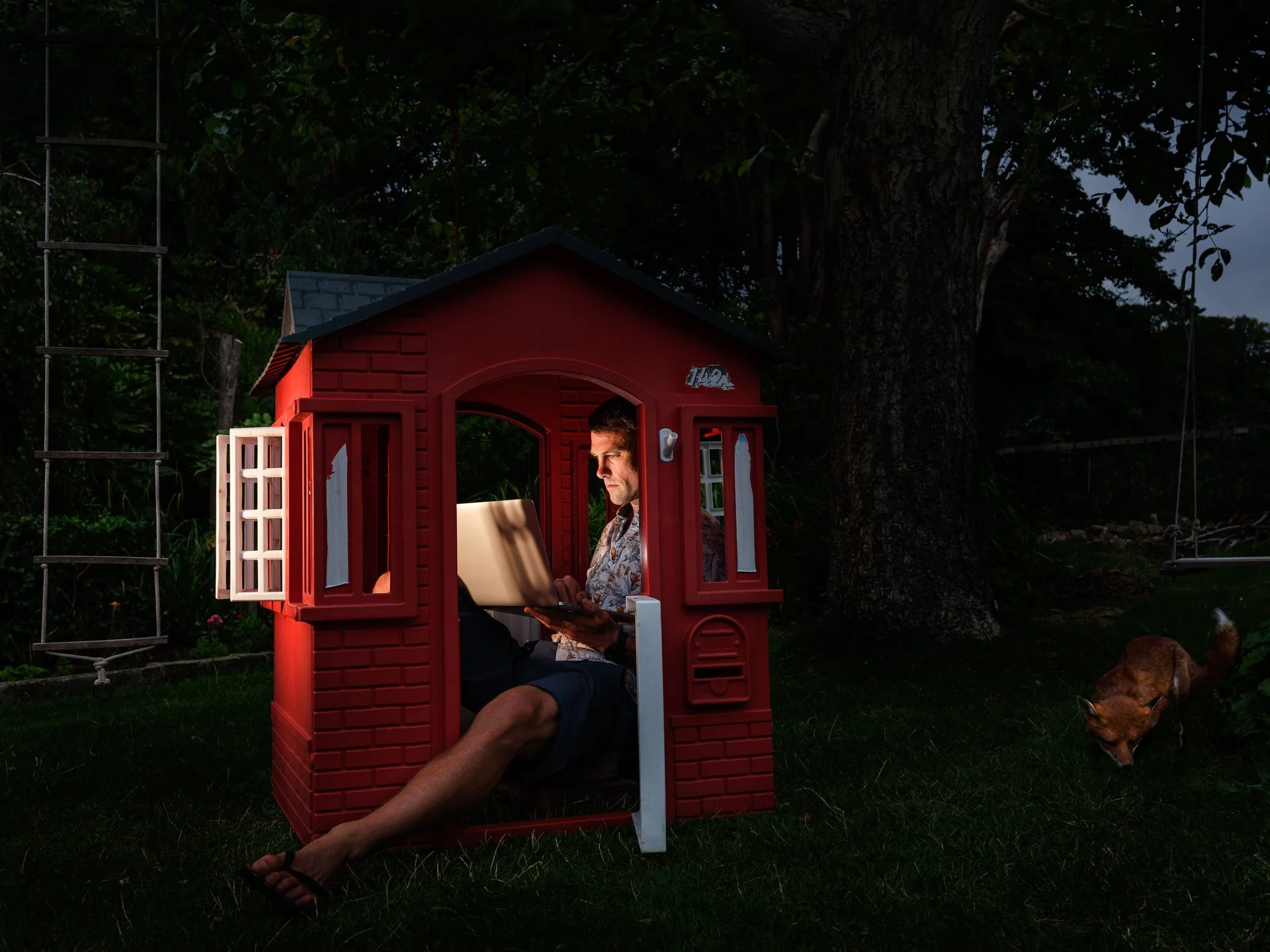 A man sitting inside a red playhouse shaped like a house, working on a laptop, while a dog is lying on the grass outside, under a large tree, during evening.