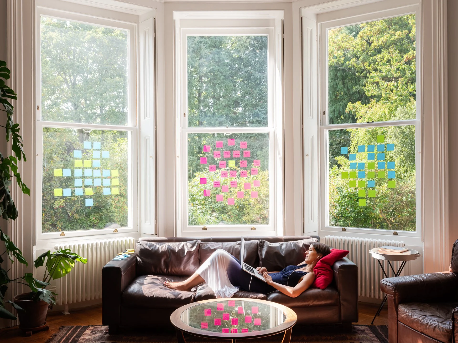 Woman relaxing on a black leather couch in a living room, using a laptop, with large windows showing green trees outside. Post-it notes are on the windows.