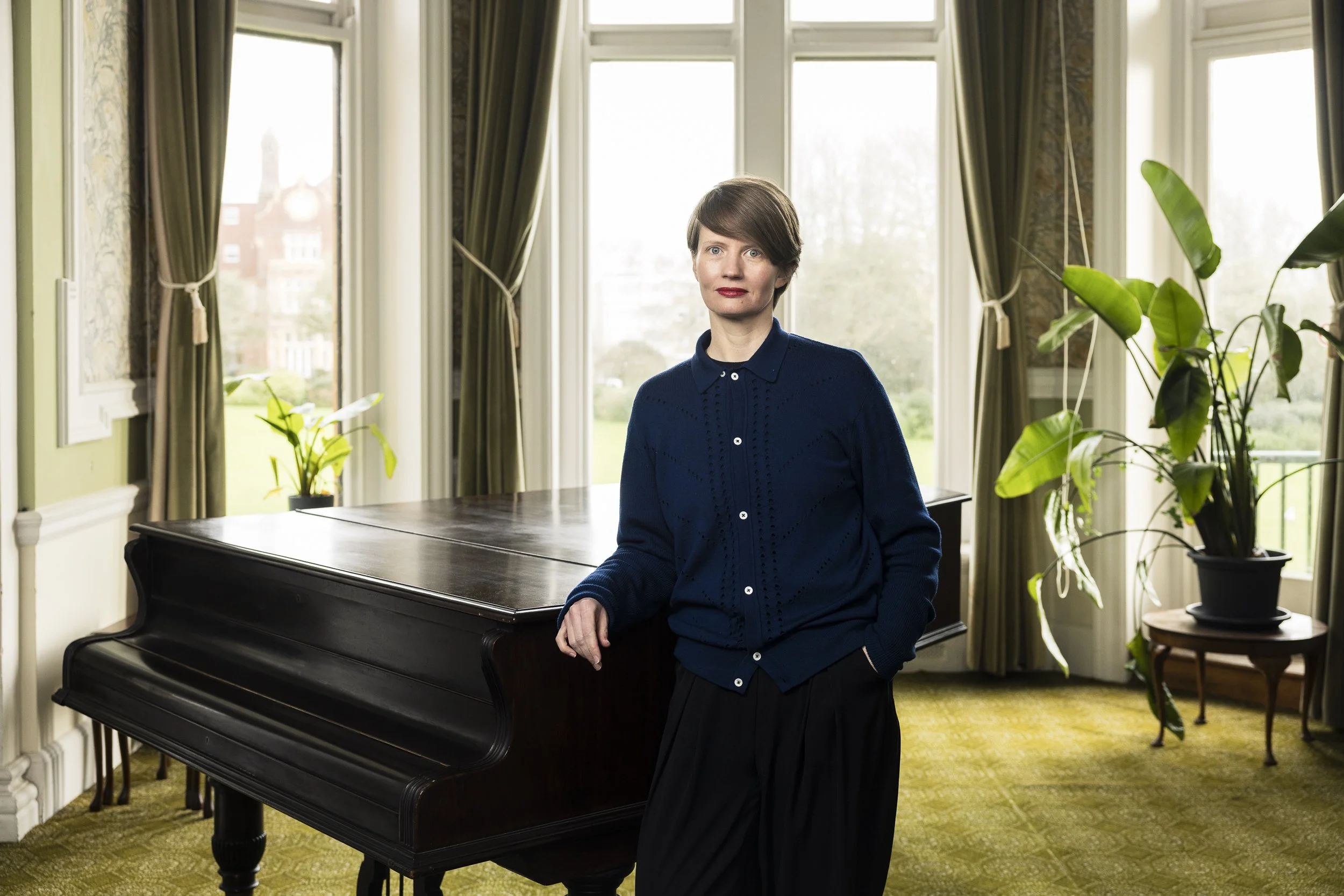 Woman with short hair and red lipstick sitting next to a black piano in a bright room with large windows, green curtains, and indoor plants.