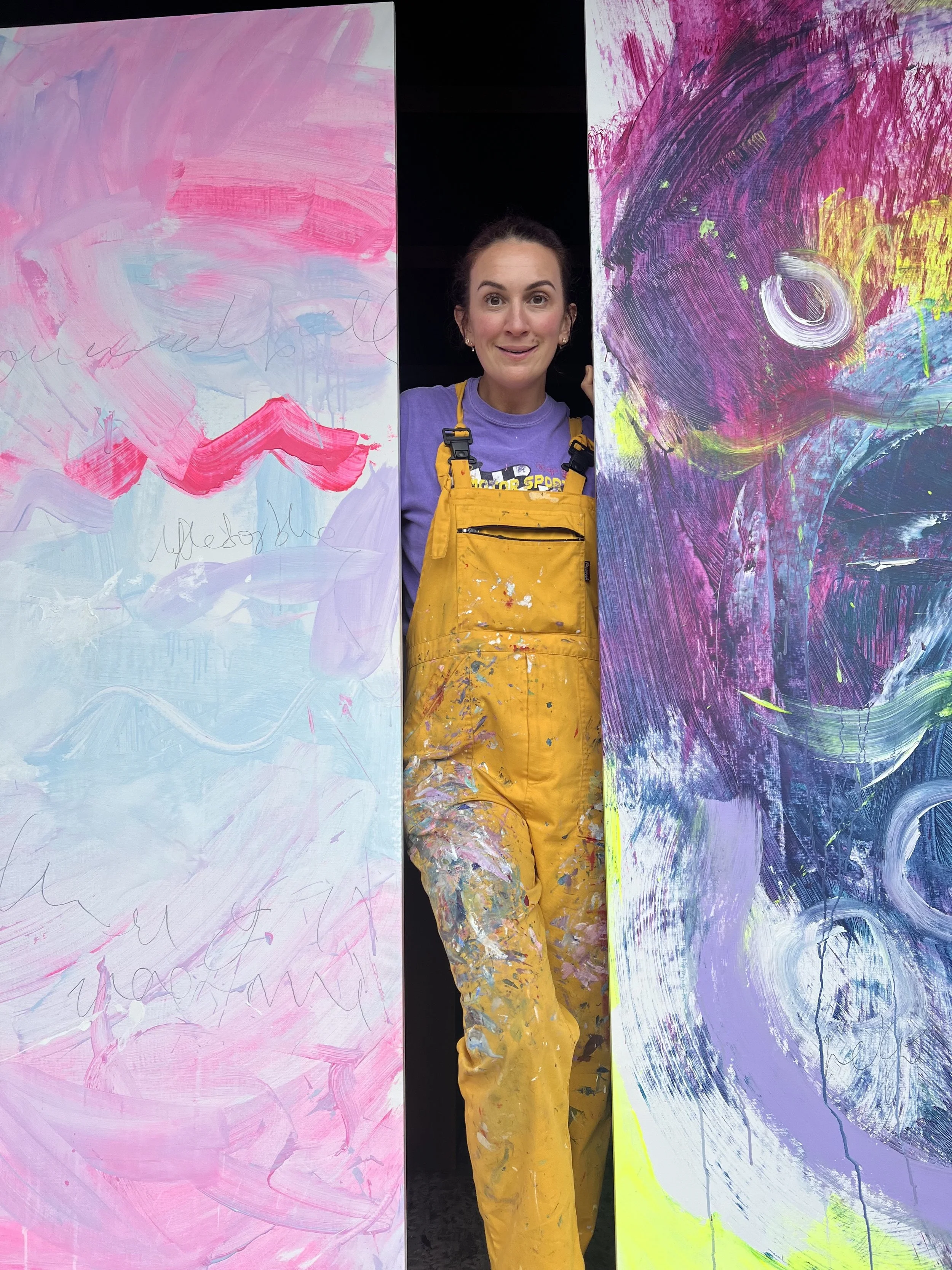 Aly Harte in yellow painting overalls standing between two large abstract paintings