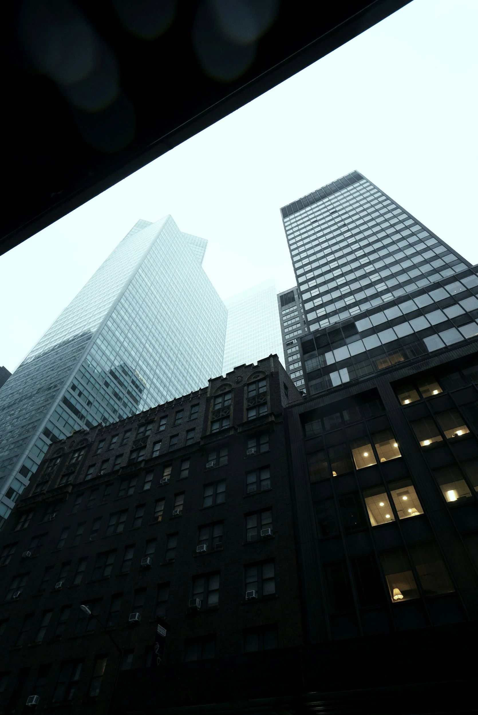 Low-angle view of tall skyscrapers in a city, viewed from a window or beneath an overhang on an overcast day.