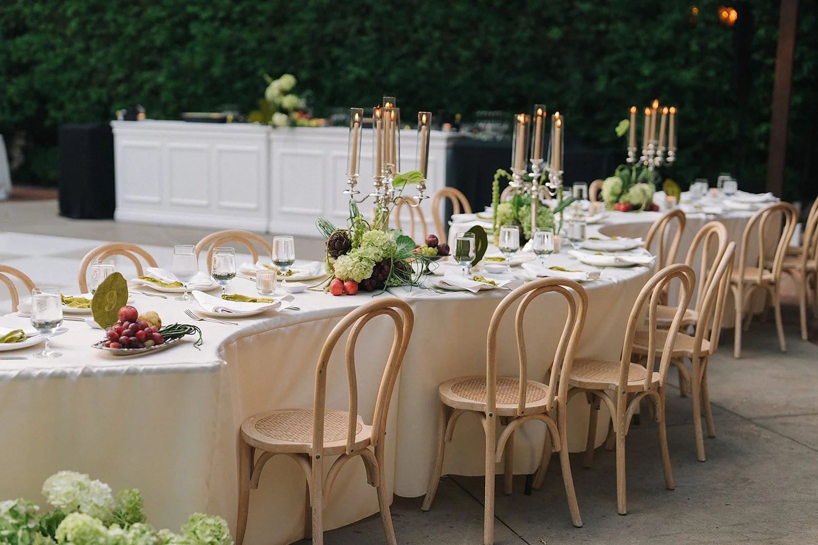 An outdoor wedding reception setup with round and rectangular tables decorated with floral centerpieces and pastel-colored table runners, surrounded by wooden chairs, with trees and mountains in the background and string lights overhead under sunny weather.