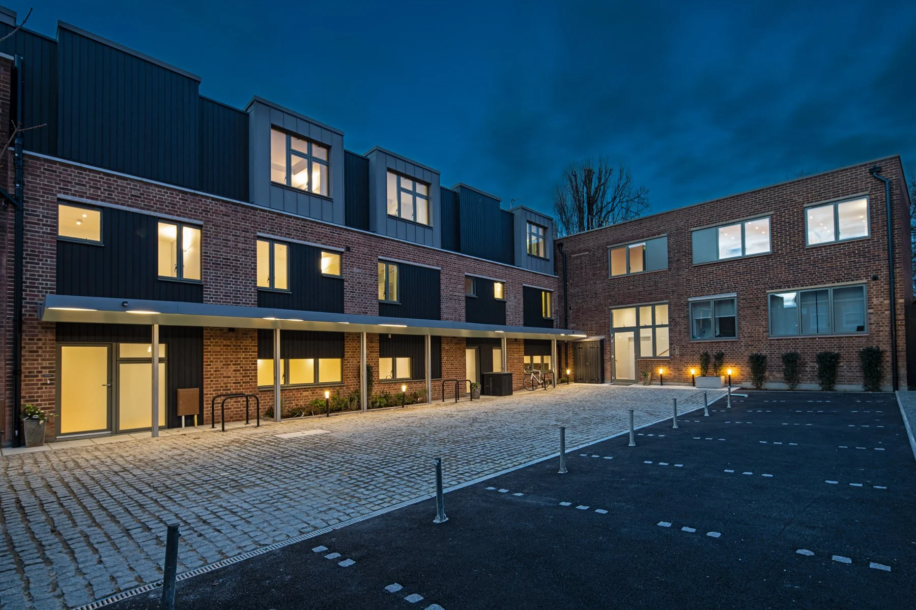 Square Feet Architects — Modern residential building at dusk with lit windows and an empty parking lot in front.