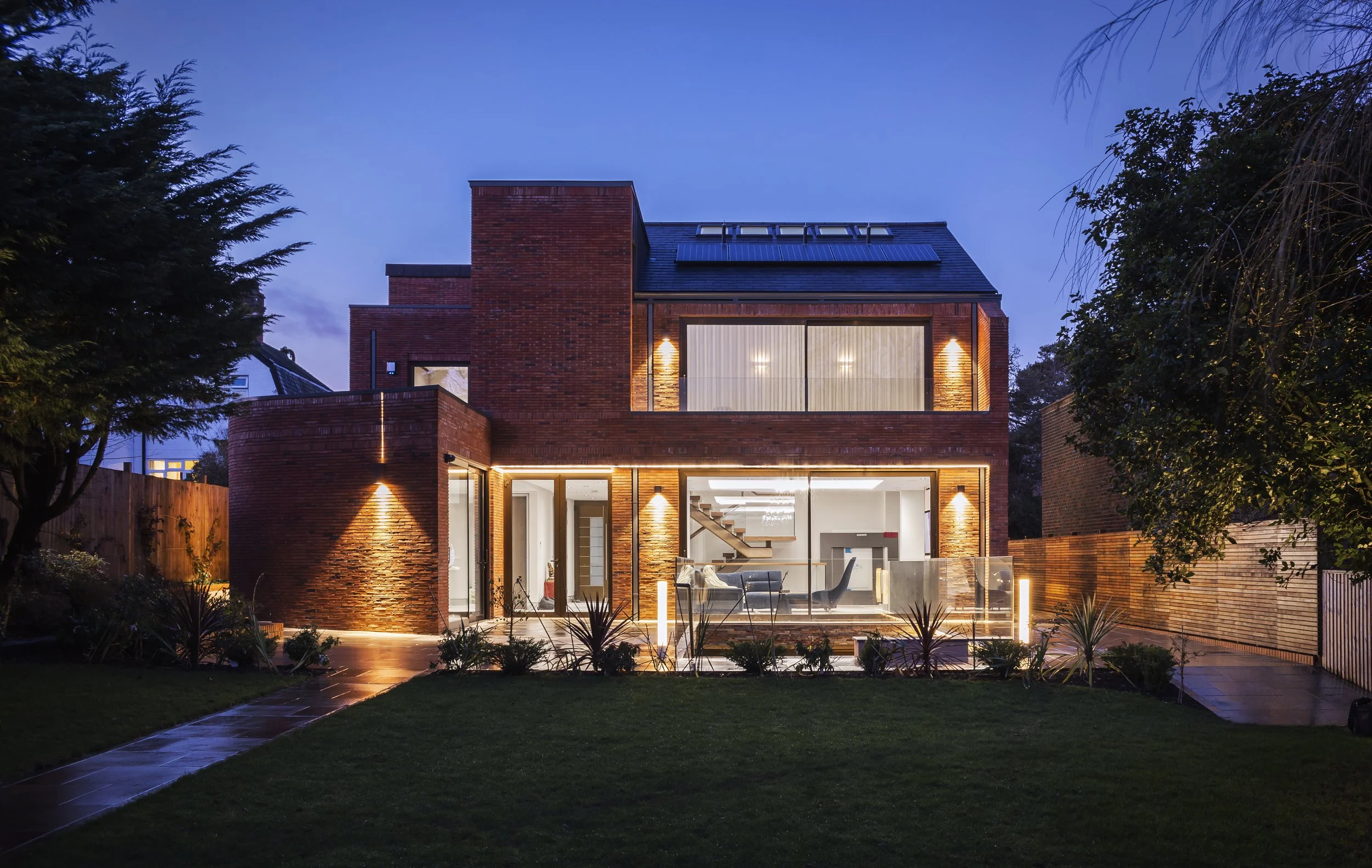 Square Feet Architects — Modern brick house illuminated at night with large glass windows and a well-maintained front yard.