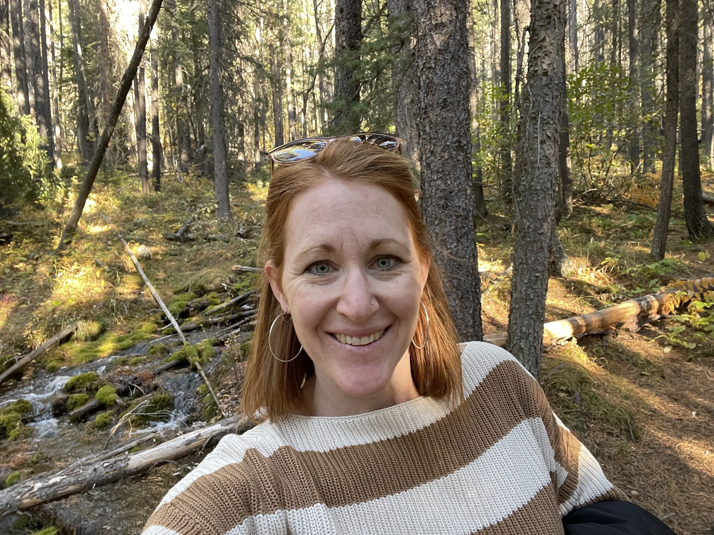 A woman with red hair smiling in a forest with tall trees, sunlight filtering through the branches, and a small stream in the background.