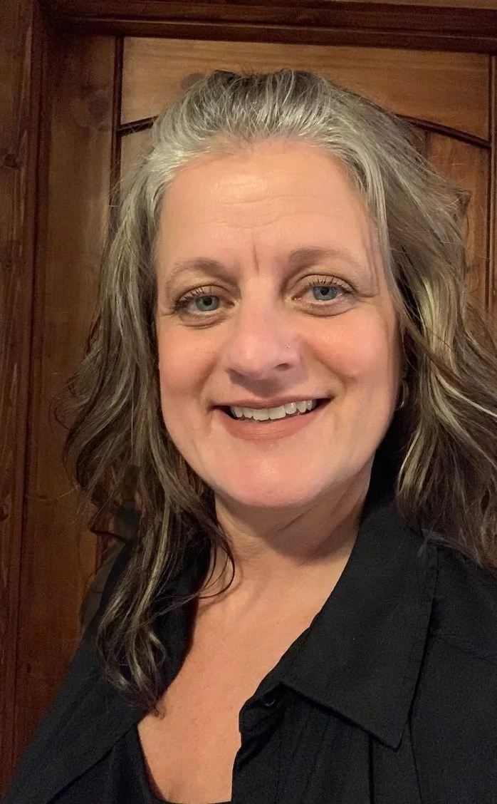 Person smiling in front of wood panel background, wearing a plaid shirt.