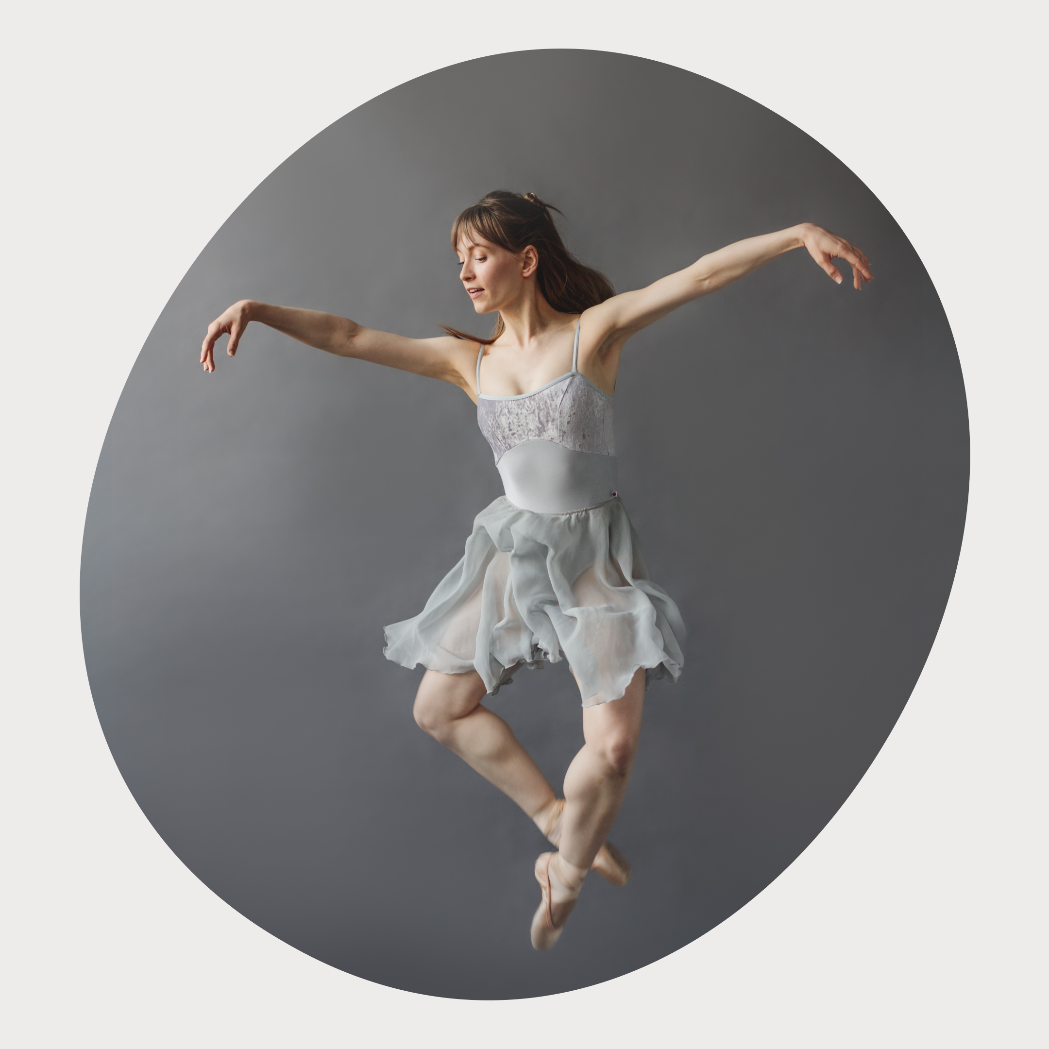 Danseuse de ballet en plein saut, effectuant un mouvement gracieux, vêtue d'une robe en mousseline gris clair avec des détails en dentelle, sur un fond gris neutre.