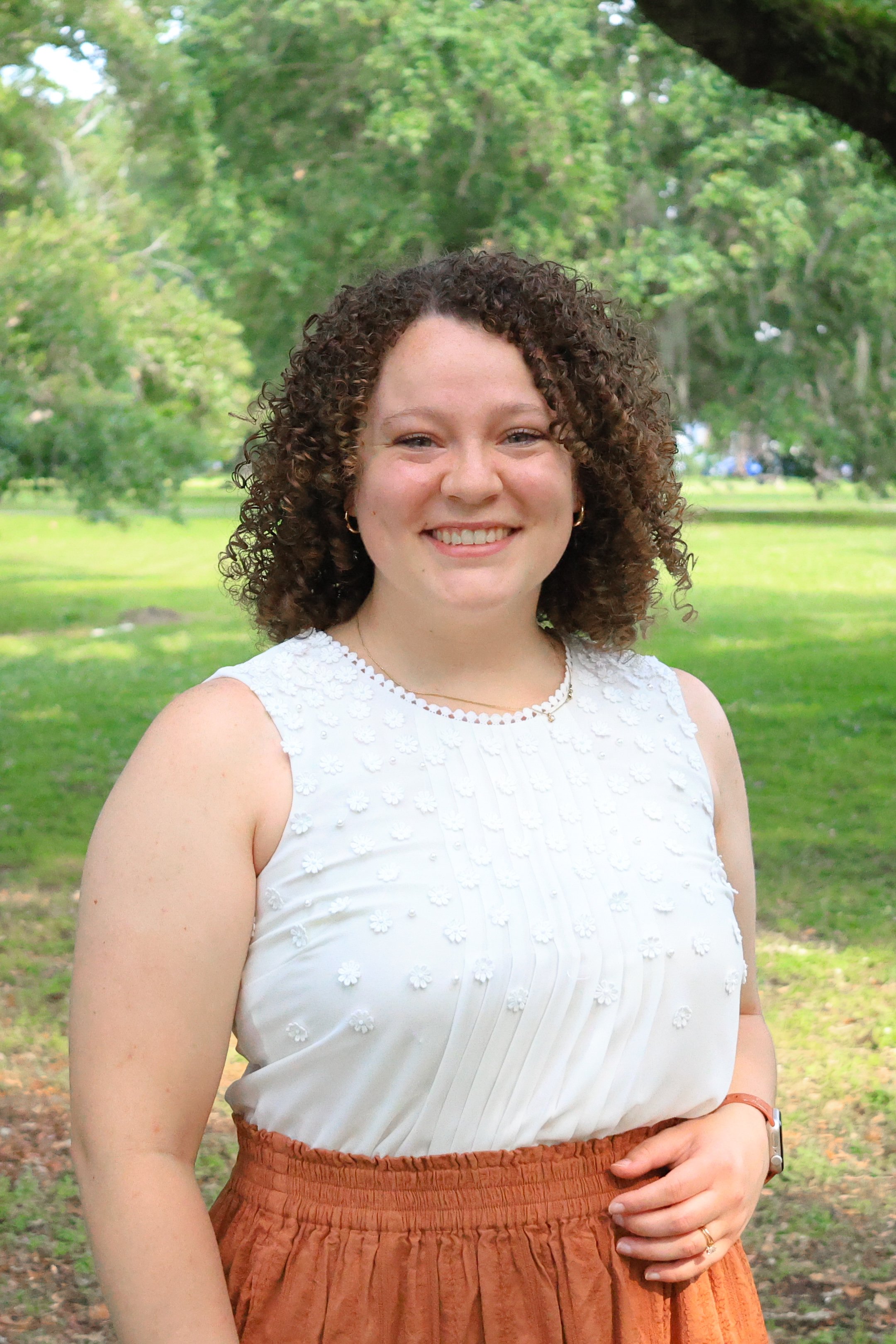 Kandis stands smiling with one hand on her hip outside in the park.