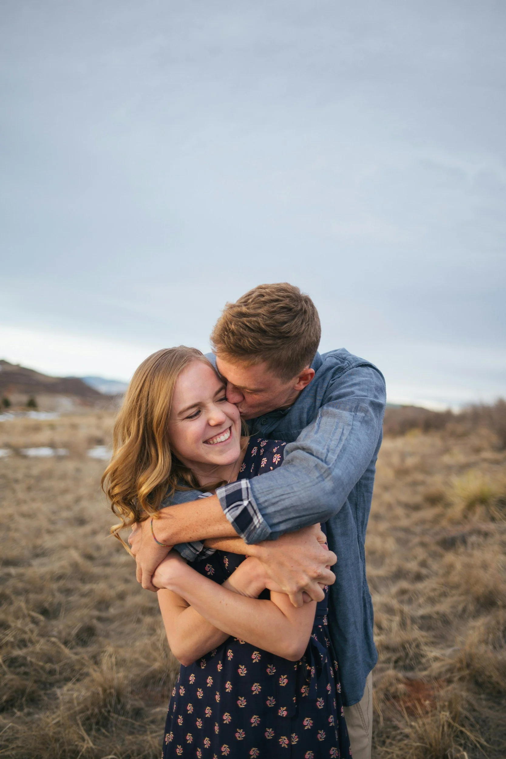 A couple hugging outdoors in a field with a cloudy sky in the background, smiling and showing affection.