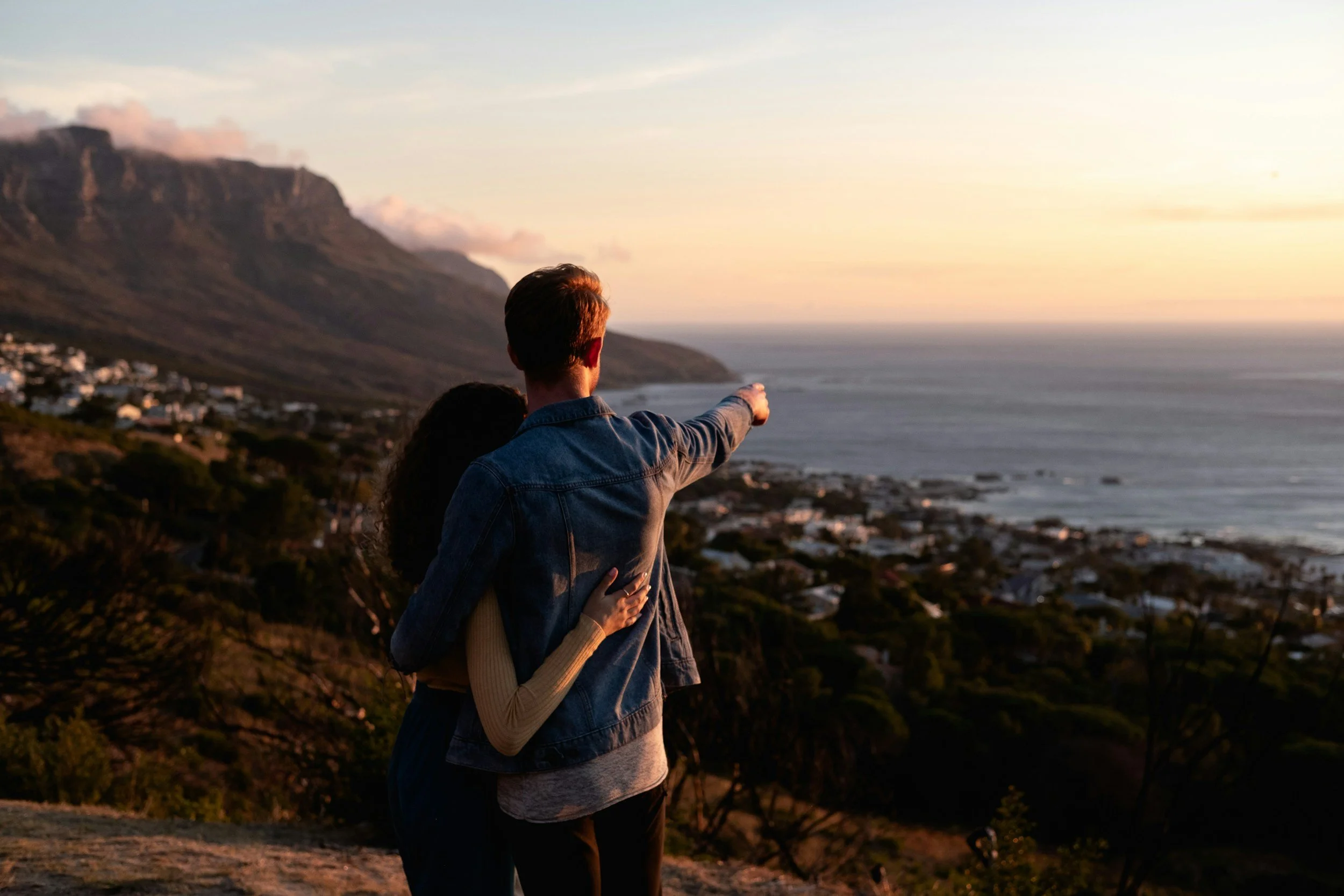 A couple embracing while standing on a hill overlooking a coastal town at sunset, with a mountain in the background.