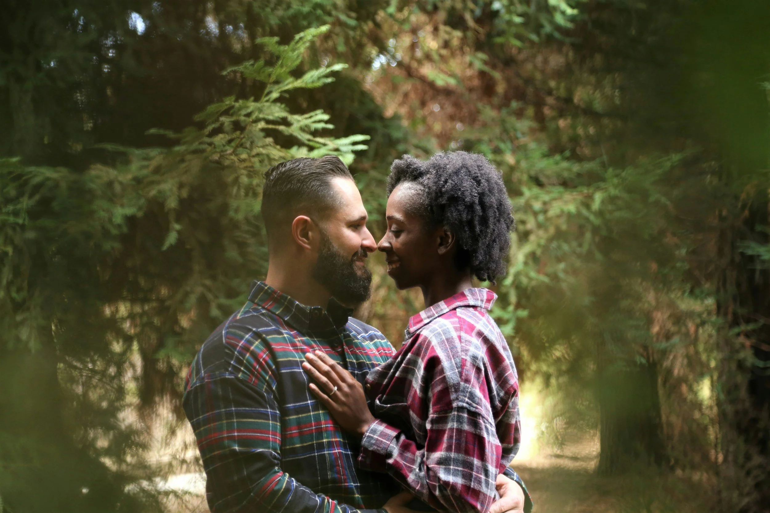 A white man and a black woman, both in plaid shirts, stand facing each other smiling with their noses touching. Green trees in the background.