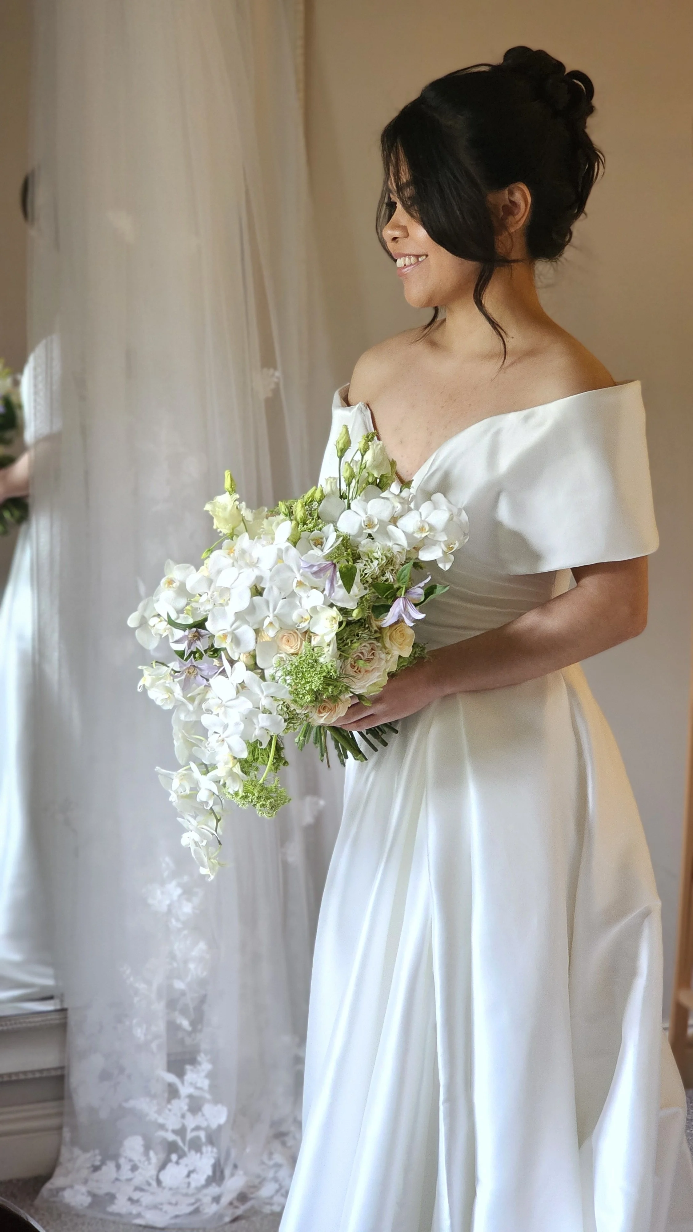 A bride in a white wedding dress holding a bouquet of white and pastel flowers, standing near a window with sheer curtains.