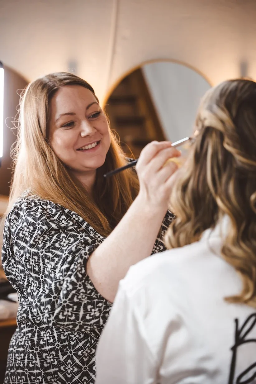 A makeup artist is applying makeup to a woman with wavy hair in a salon or studio setting.