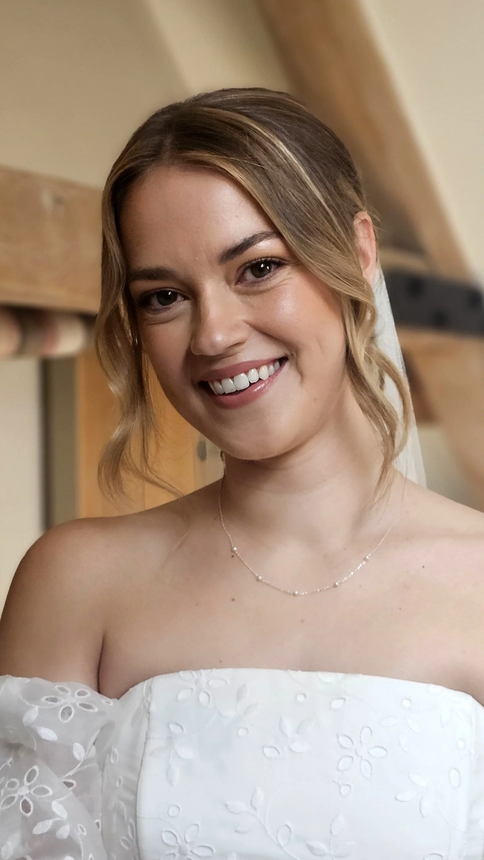 A woman smiling at the camera, wearing a white off-shoulder dress with floral embroidery and a delicate necklace, in an indoor setting with wood beams in the background.
