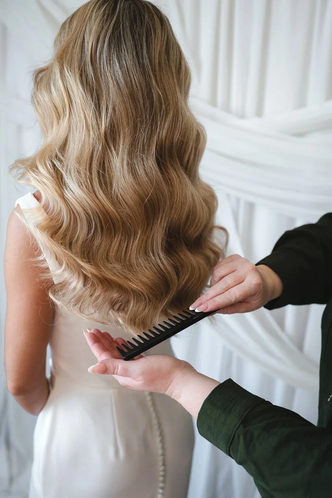 Close-up of a person holding a black comb, styling long, wavy, blonde hair on a woman wearing a white dress, with white drapery in the background.