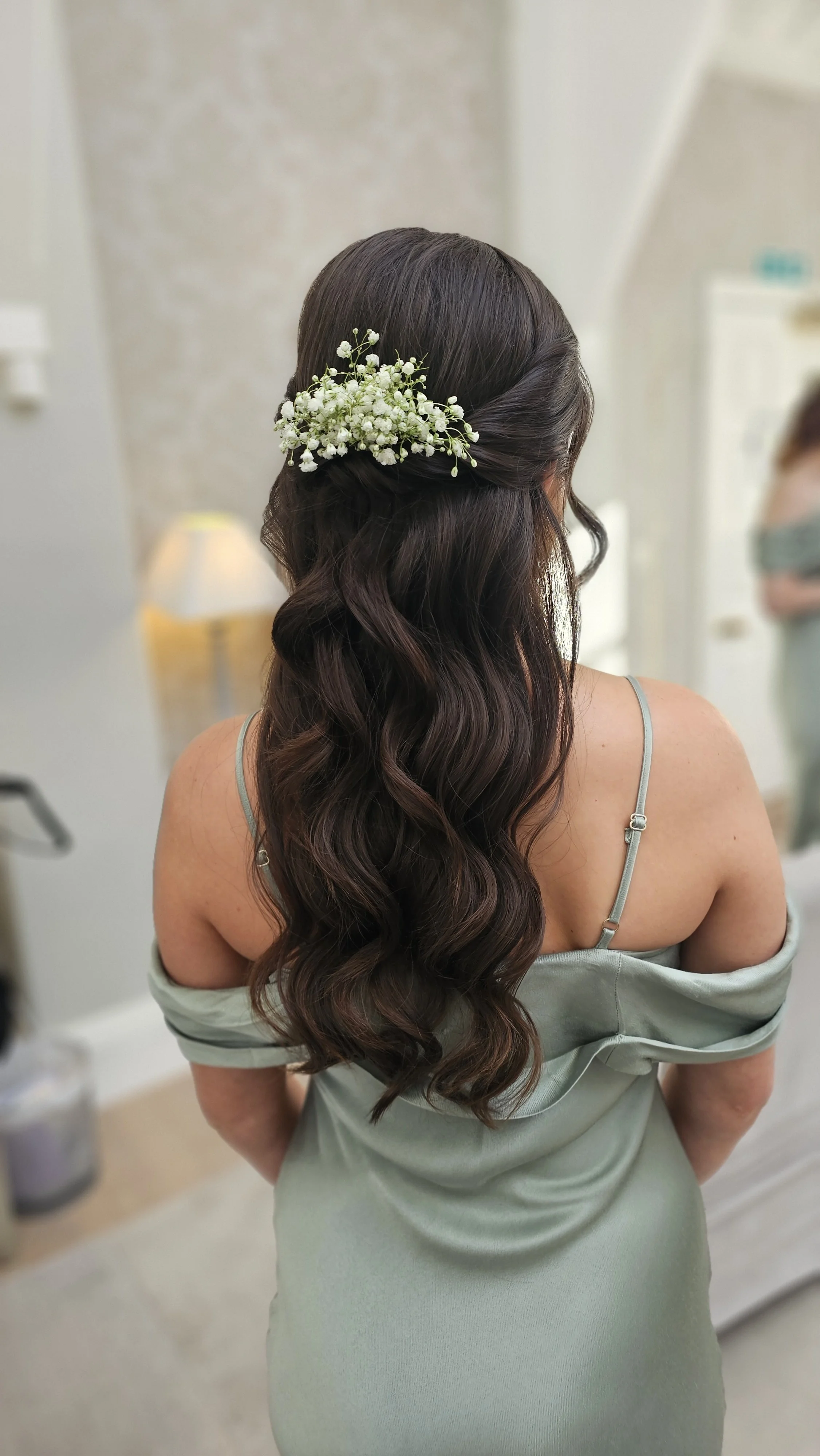 A woman with long wavy brown hair adorned with a small bouquet of white flowers, wearing a light green off-shoulder dress with spaghetti straps, standing in a room with blurred background.