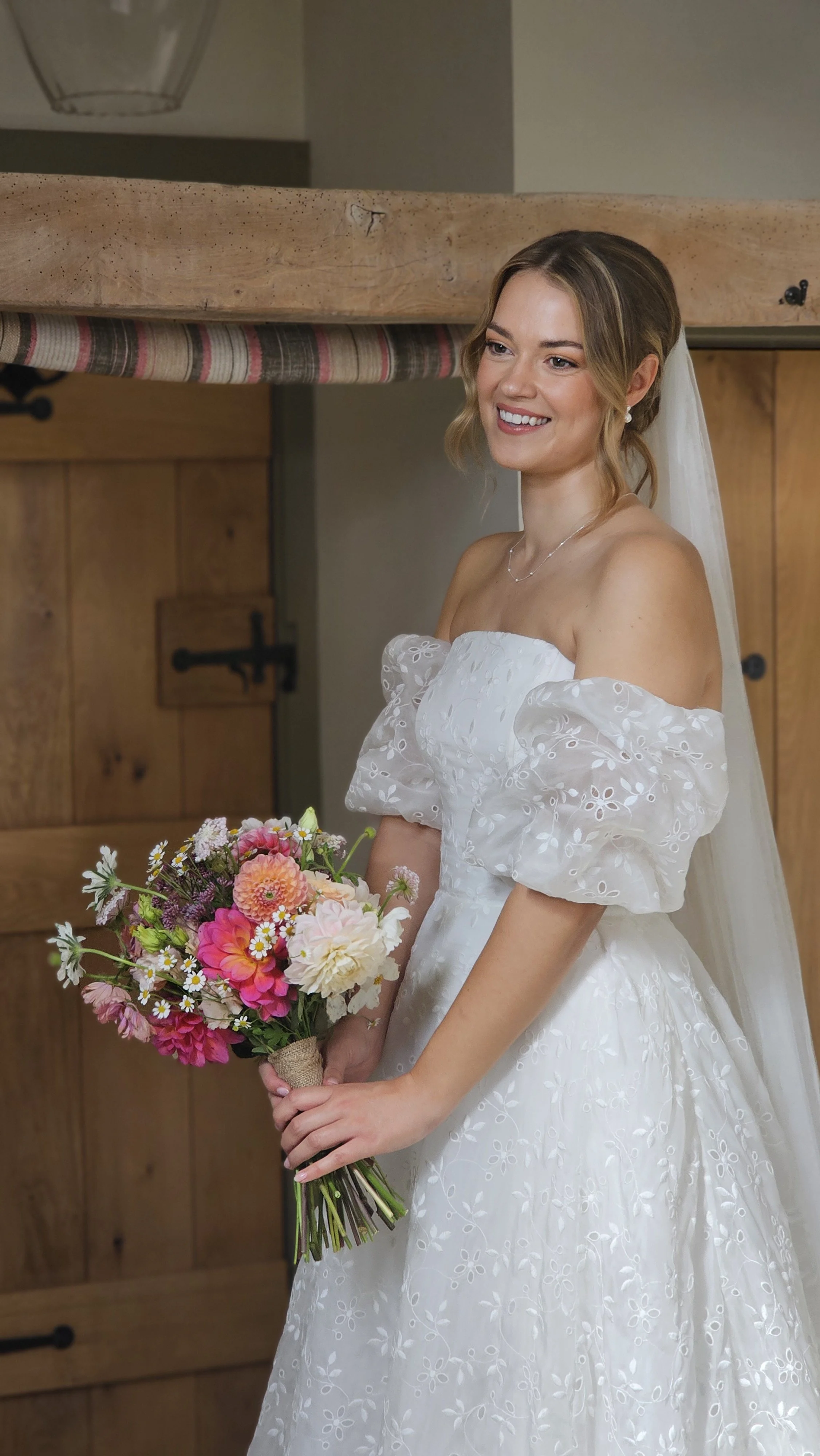 A smiling bride in a strapless white wedding dress with floral embroidery, holding a bouquet of pink and white flowers, standing indoors in front of wooden doors.