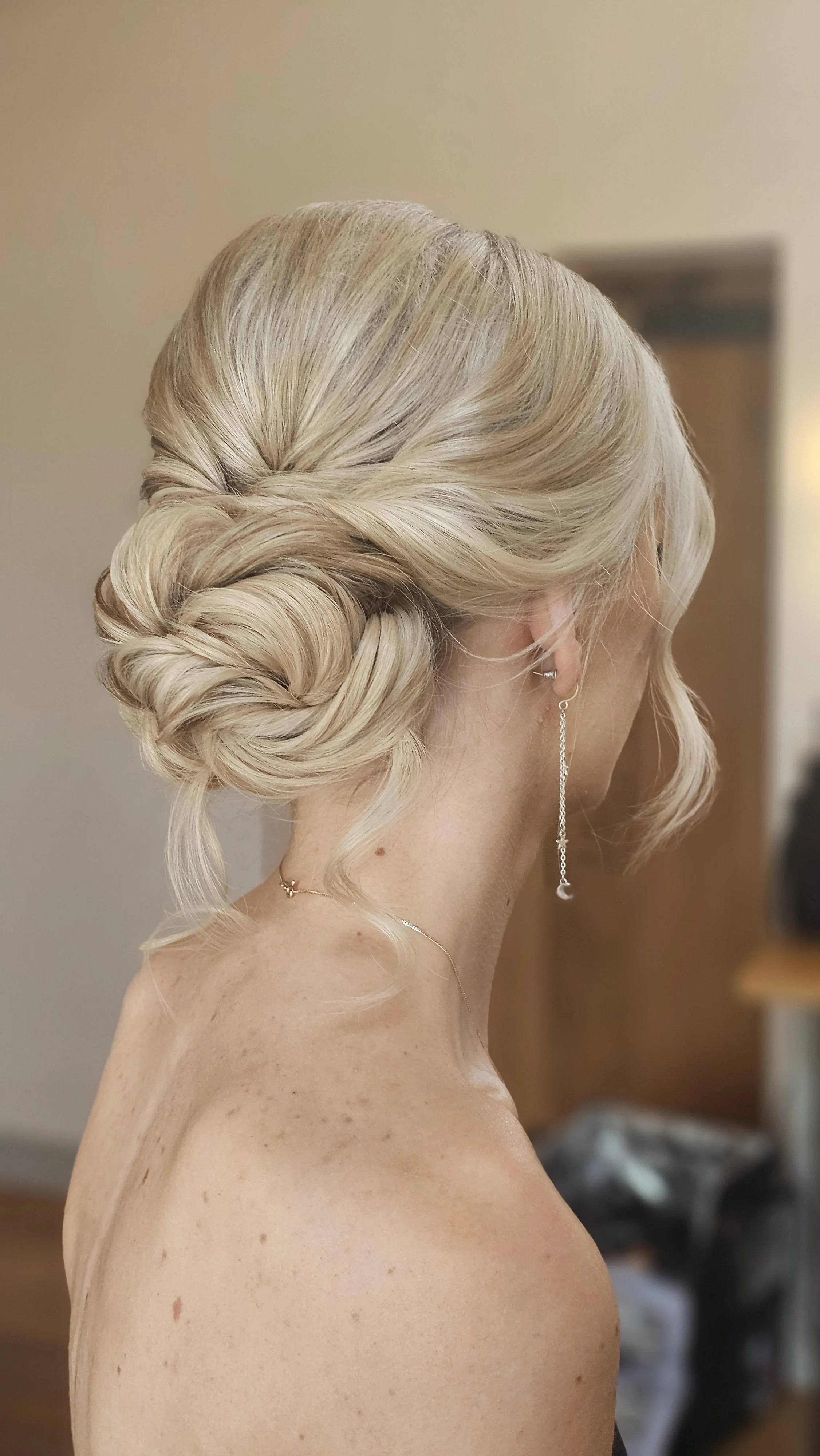 A woman with blonde hair styled in an elegant updo, wearing long dangling earrings and a delicate necklace, in an indoor setting with a blurred background.