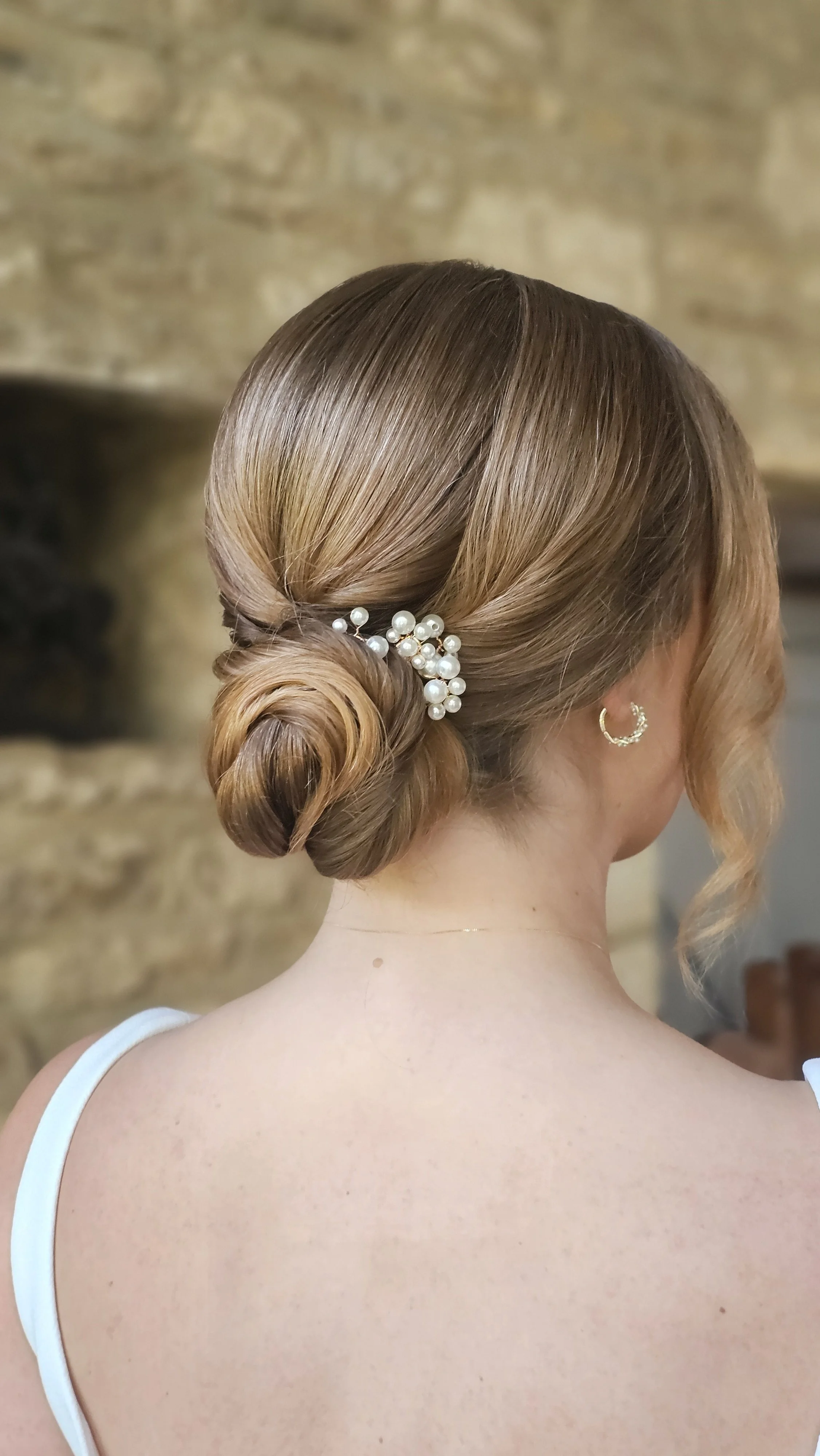 Back view of a woman with elegant updo hairstyle adorned with pearl hairpin and wearing gold hoop earrings, with a blurred stone wall background.
