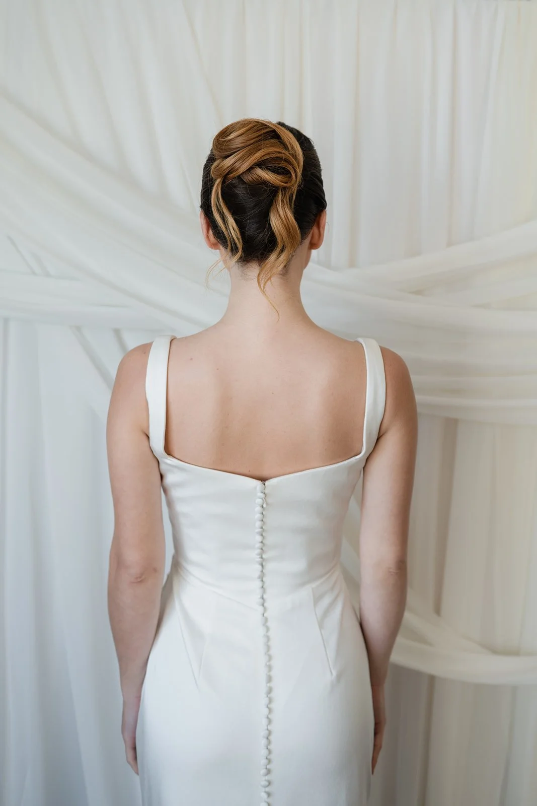 Back view of a woman in a white wedding dress with a button closure down the back, standing in front of a white fabric backdrop.