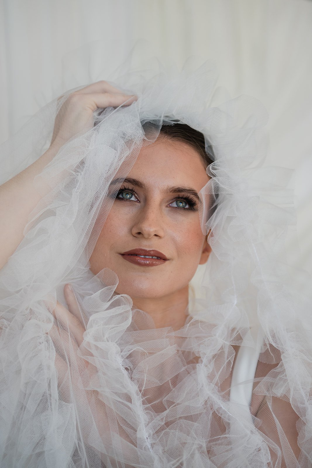 A woman with blue eyes and brown hair, wearing a white tulle veil, holding it with her hand, smiling gently.