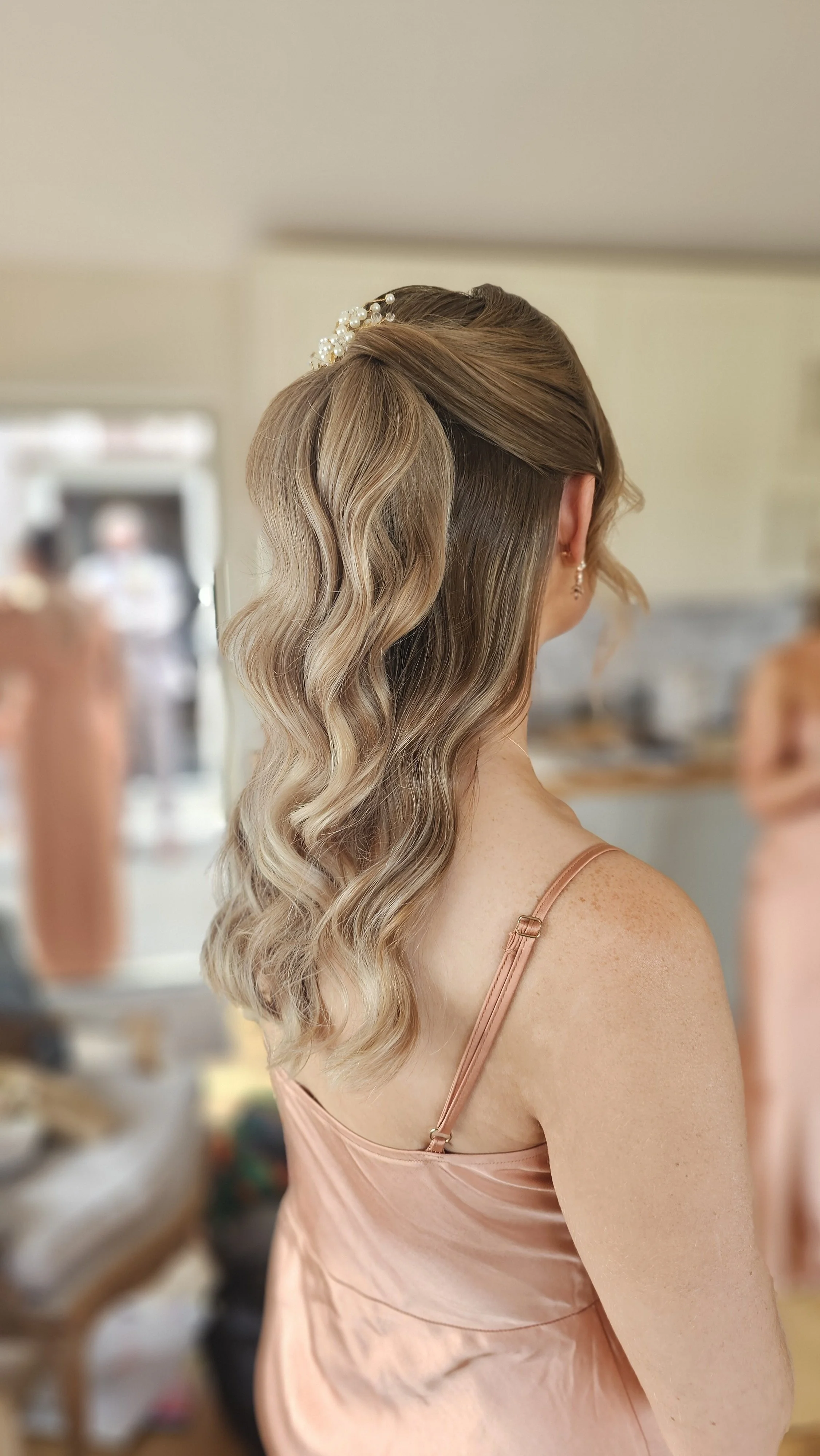 Side view of a woman with long, wavy blonde hair, wearing a peach-colored dress with spaghetti straps, adorned with a pearl hairpin, standing indoors with a blurred background.